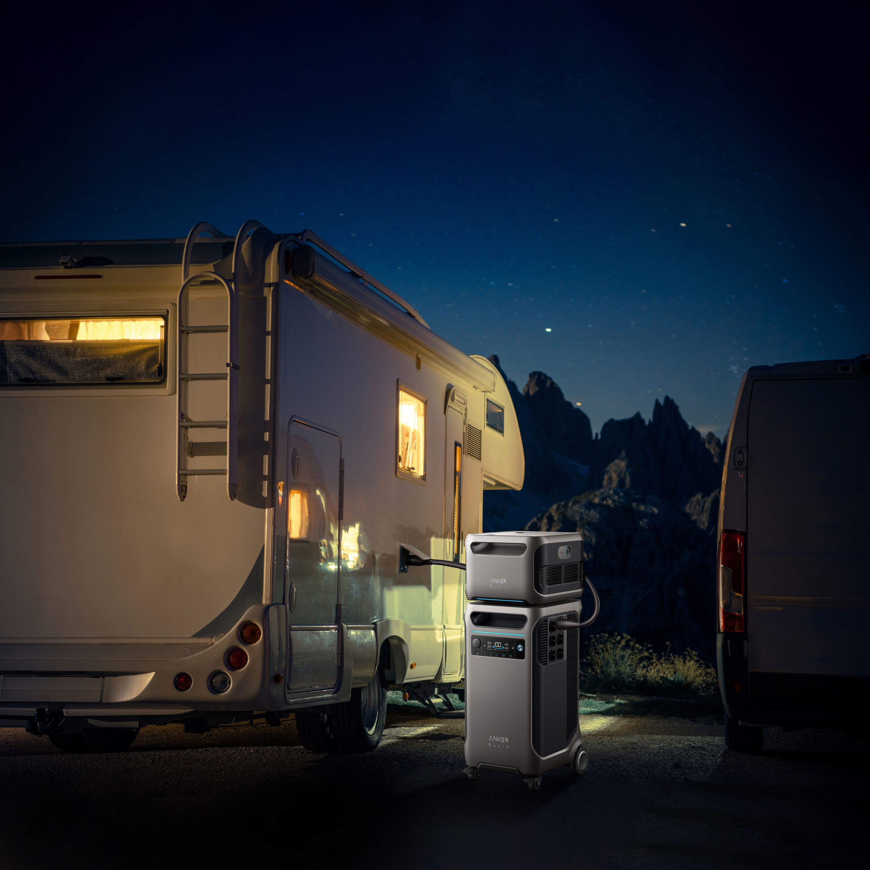 Kamper i furgon zaparkowane w nocy w pobliżu gór. Przenośna stacja zasilania stoi między nimi, podświetlona, pod gwiaździstym niebem.