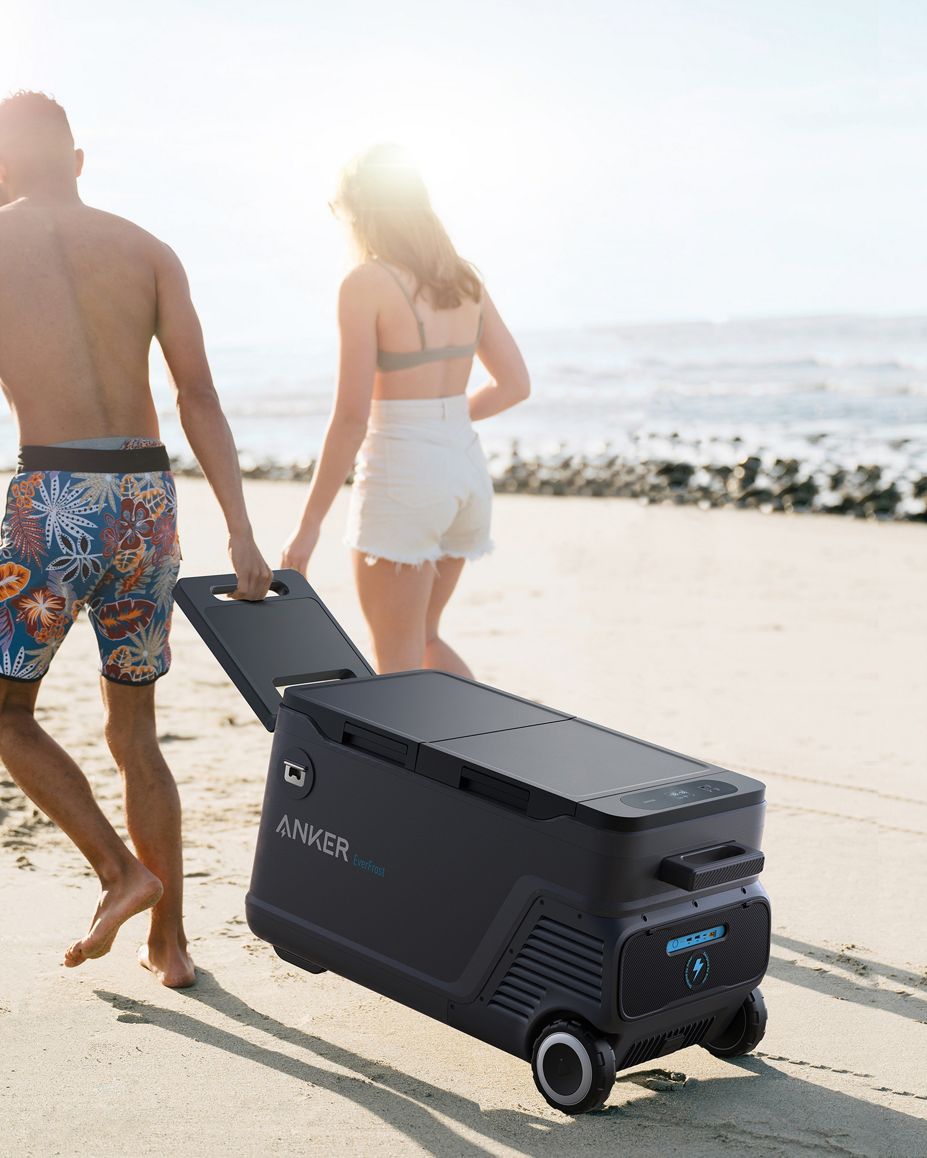 Ein Paar geht am Strand spazieren und zieht eine schwarze Kühlbox mit Rädern.