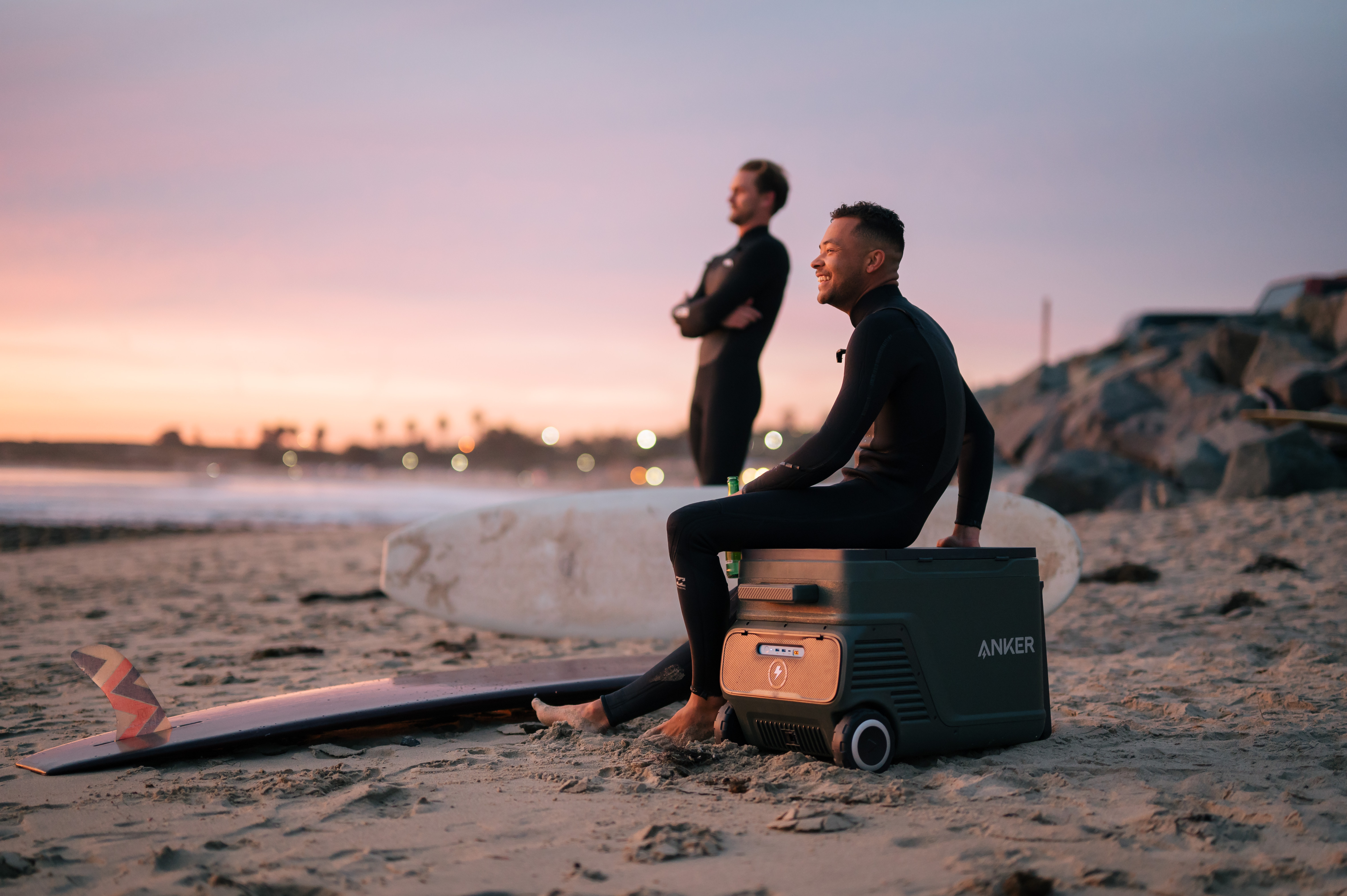 Zwei Männer am Strand bei Sonnenuntergang. Einer sitzt auf einer Kühlbox, der andere steht im Hintergrund.