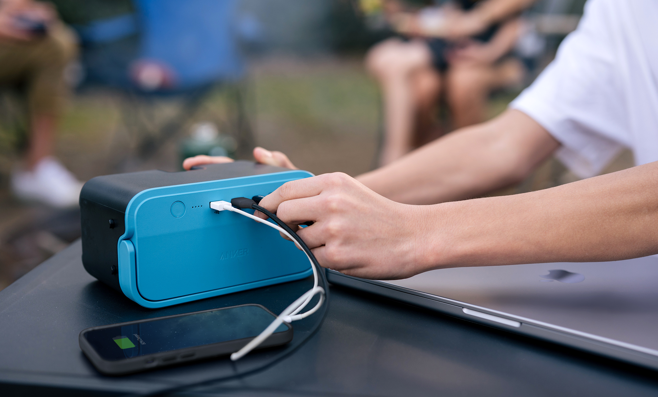 Person lädt Telefon und Laptop mit einer blau-schwarzen Powerbank auf einem Tisch im Freien.