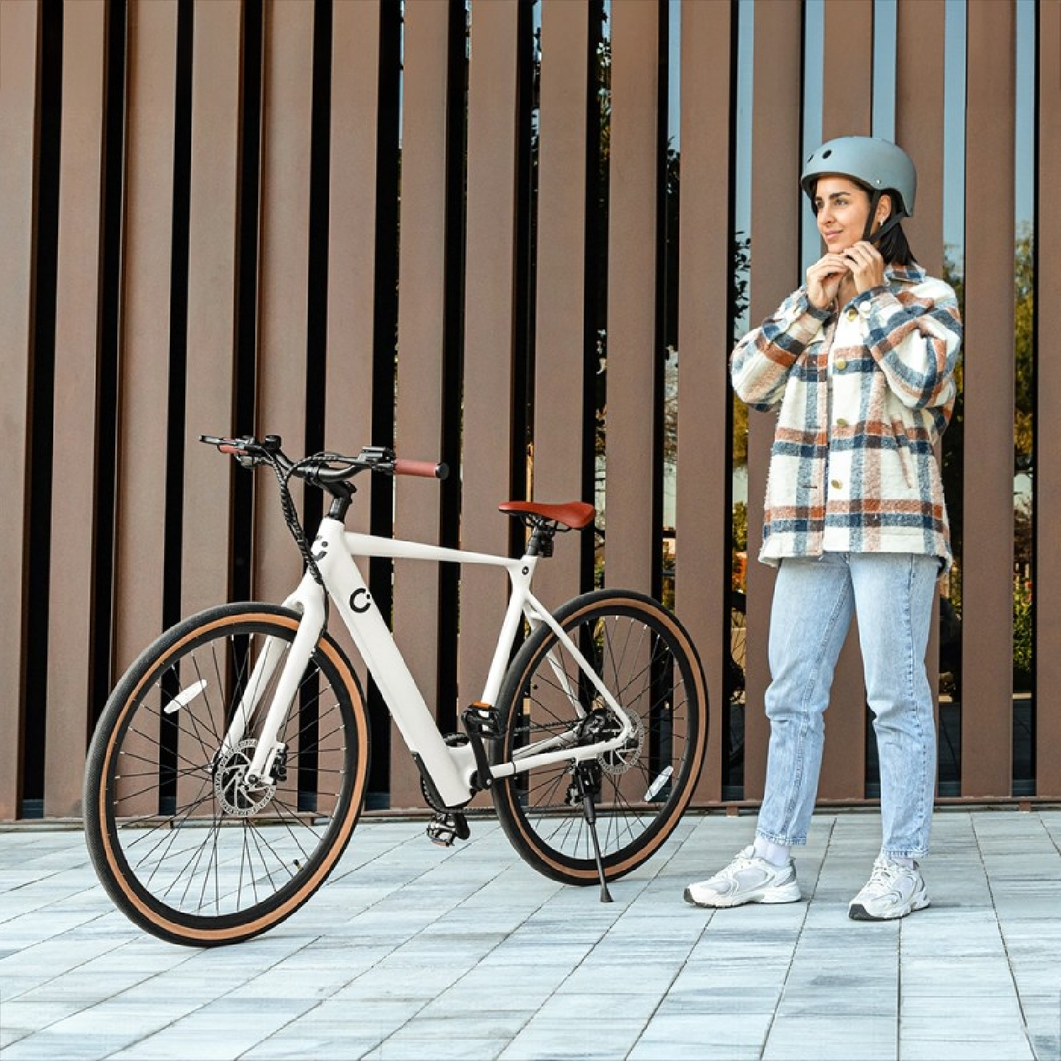 Una mujer con casco junto a una bicicleta blanca. La escena es cerca de una pared marrón.