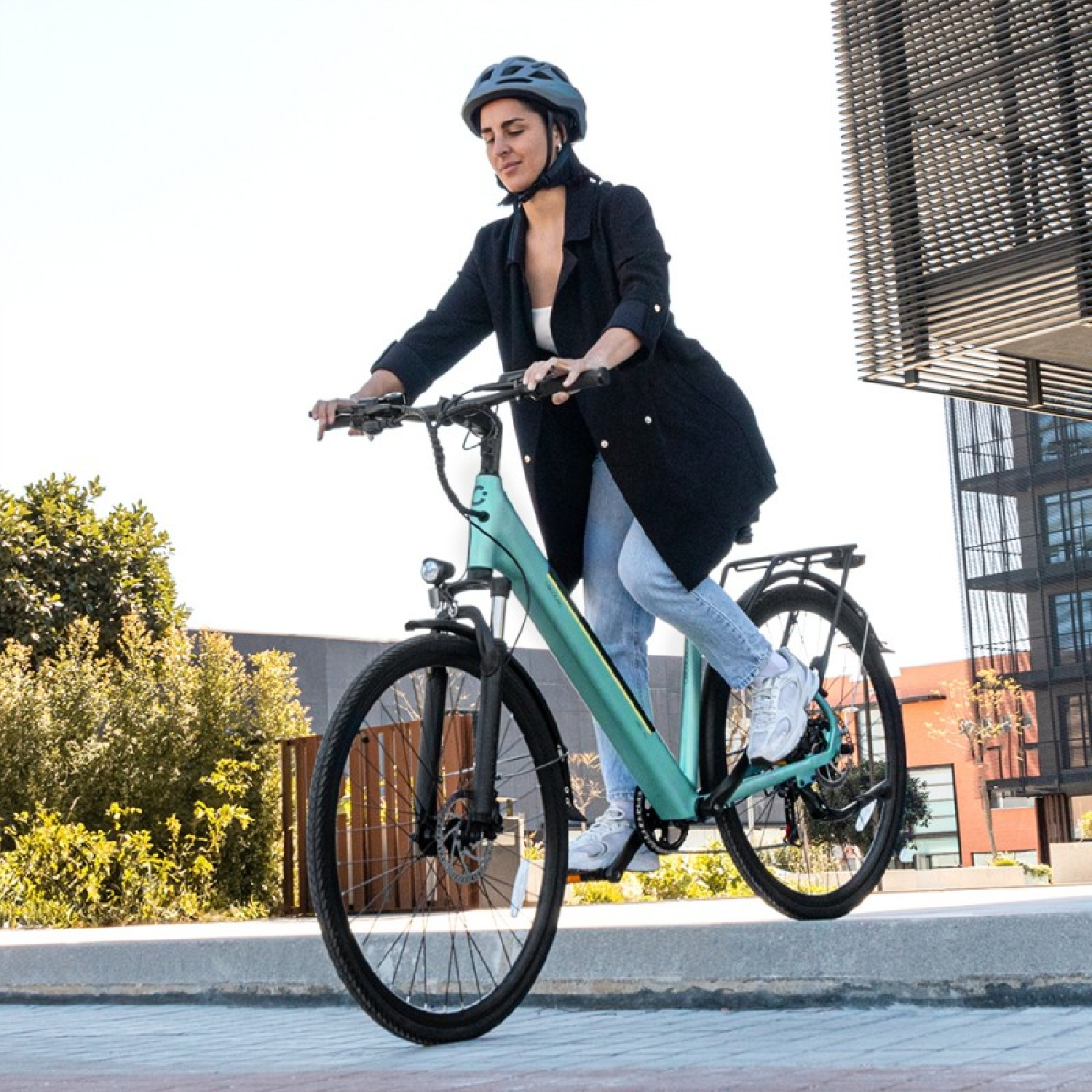 Frau fährt an einem sonnigen Tag ein türkisfarbenes E-Bike. Sie trägt einen Helm und eine Jacke.