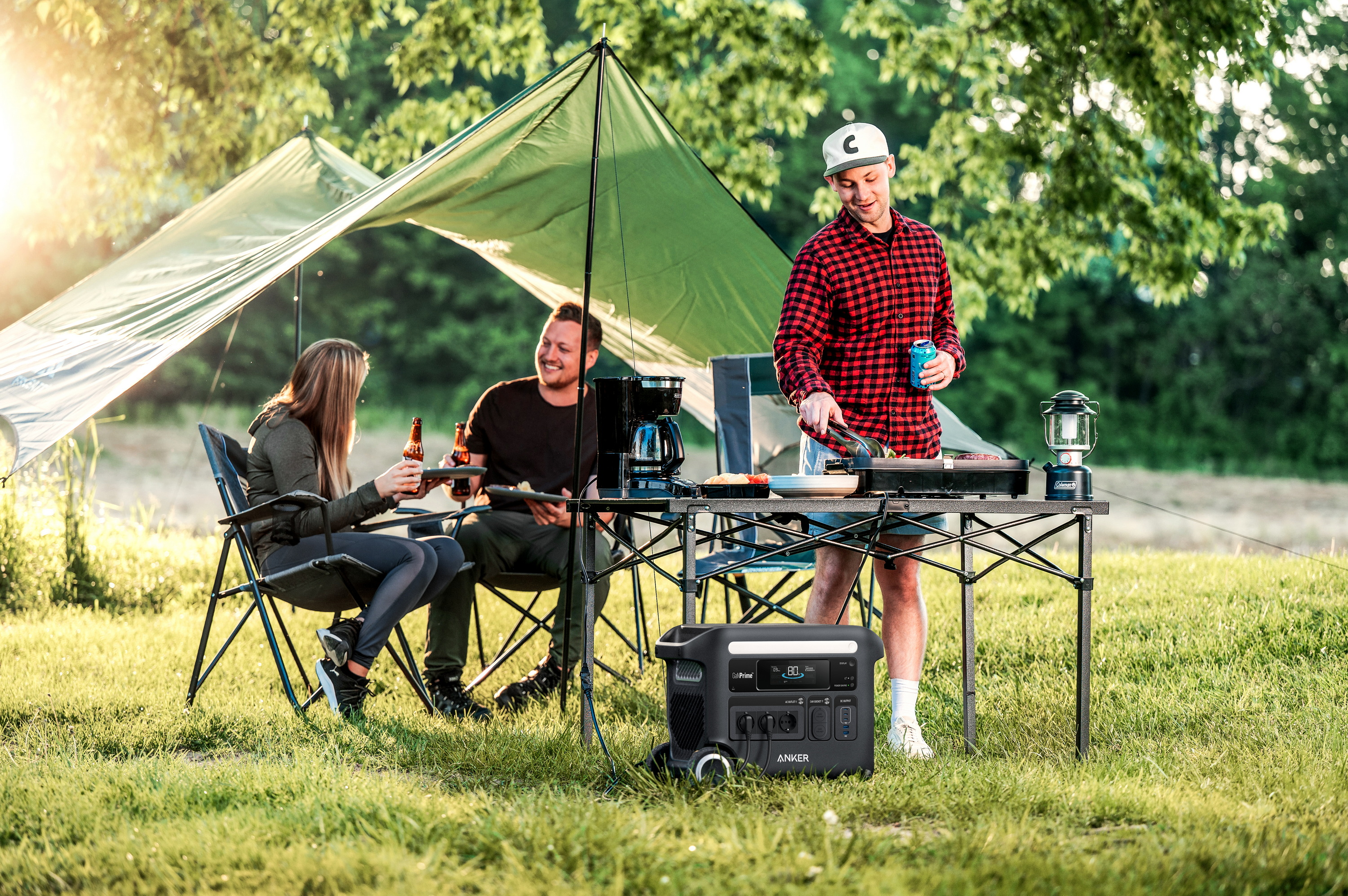 Ludzie biwakują, ciesząc się posiłkiem. Są w pobliżu namiotu, ze stołem, jedzeniem i stacją zasilania na trawiastym terenie.