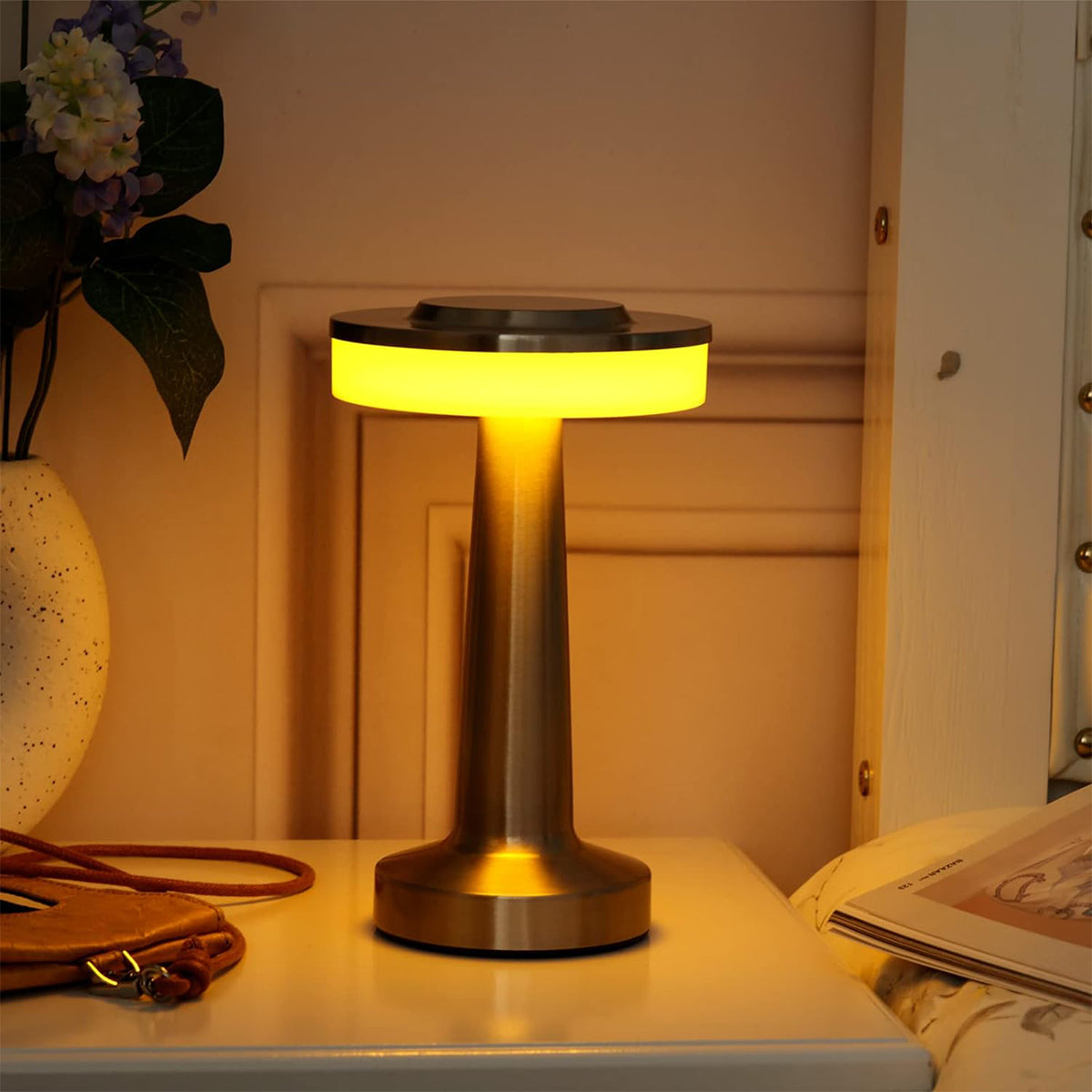 Gold-silberne Nachttischlampe mit hellgelb beleuchteter Oberseite, auf weißer Oberfläche.
