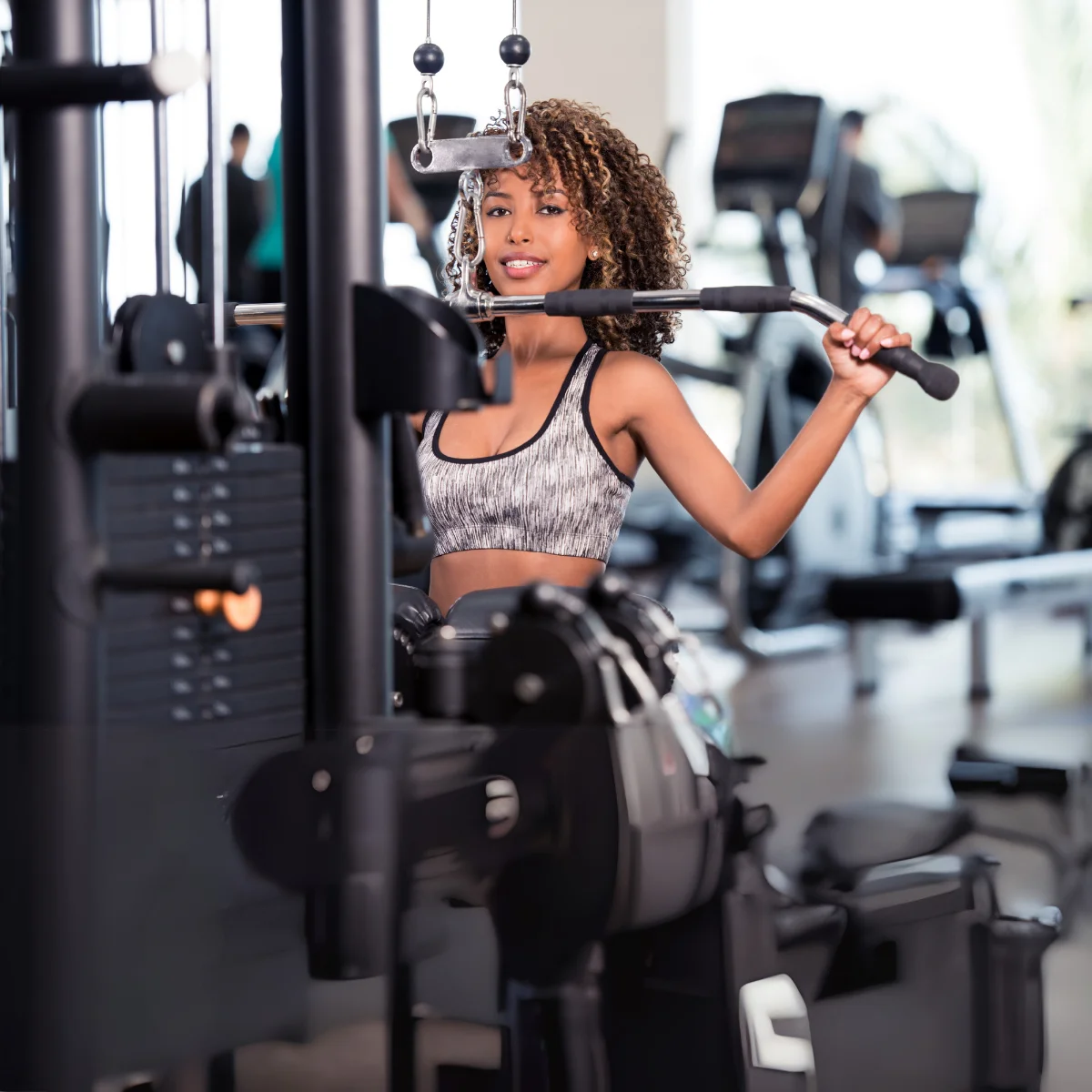 A woman using a gym machine. She is smiling. She is wearing a sports bra.