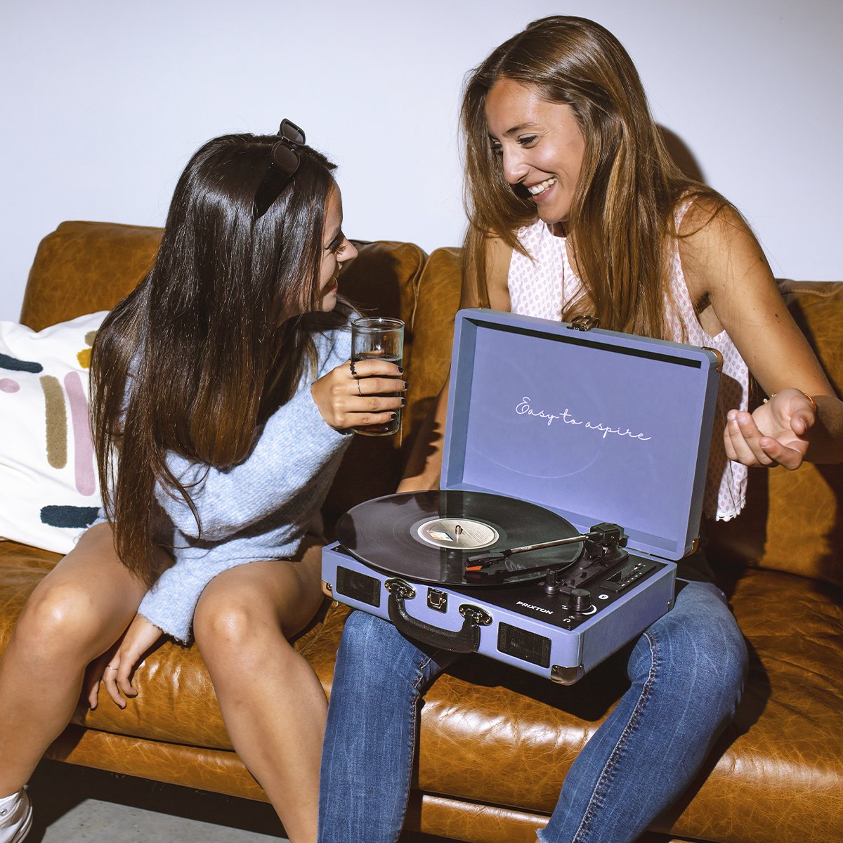 Dwie kobiety siedzące na brązowej kanapie, jedna trzymająca szklankę, w pobliżu niebieskiego gramofonu.