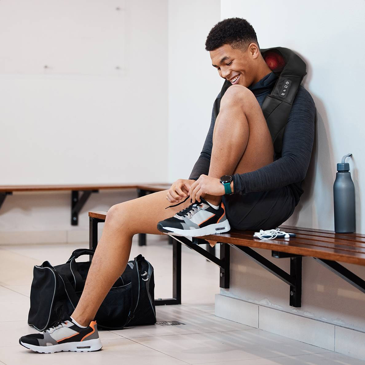 A man in a locker room, tying his shoe. He is wearing a massage device around his neck and has a bag.