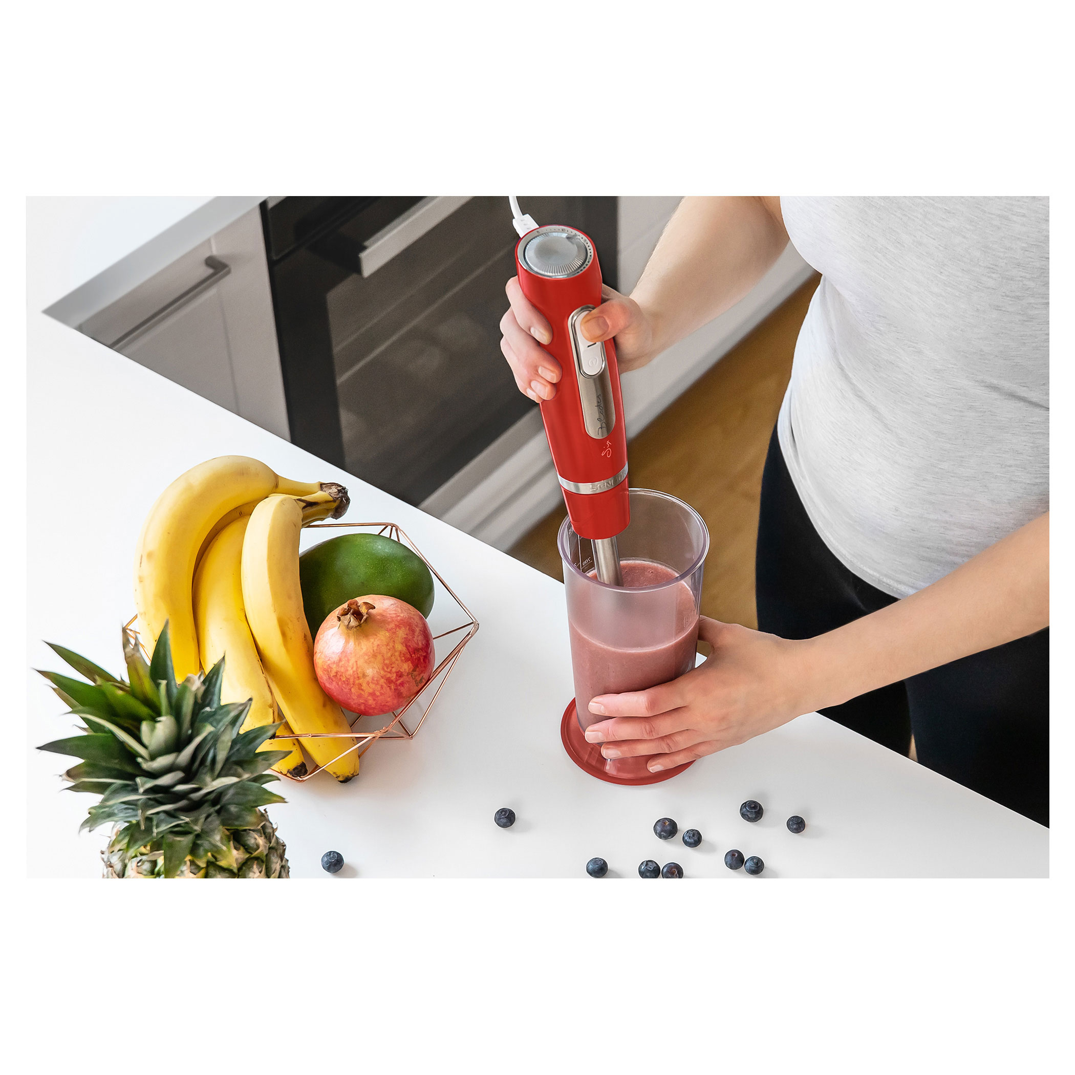 Kobieta miksuje koktajl czerwonym blenderem ręcznym. Banany, mango i granat na blacie.