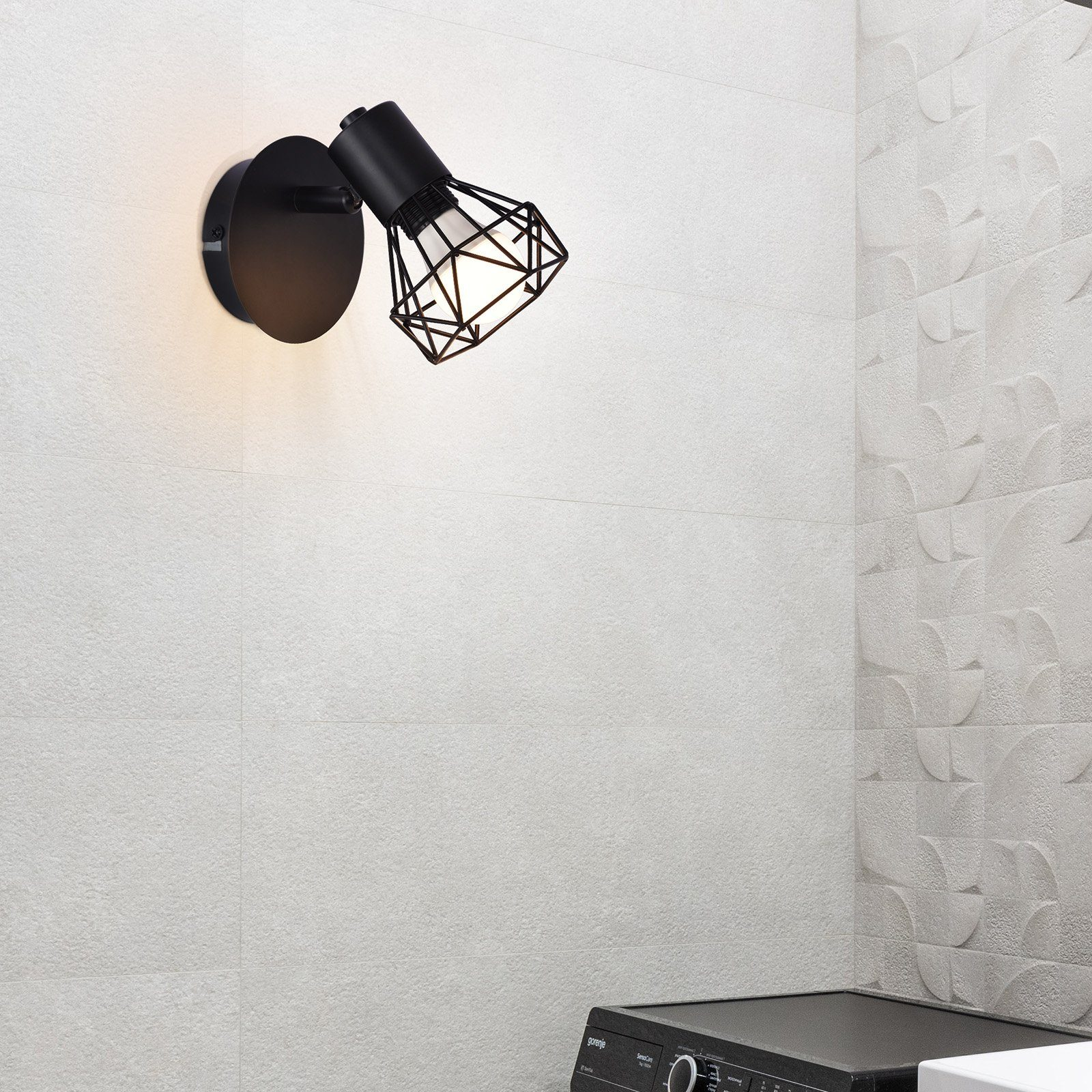 Un foco negro montado en la pared con un diseño de jaula geométrica, que ilumina una pared de azulejos blancos.