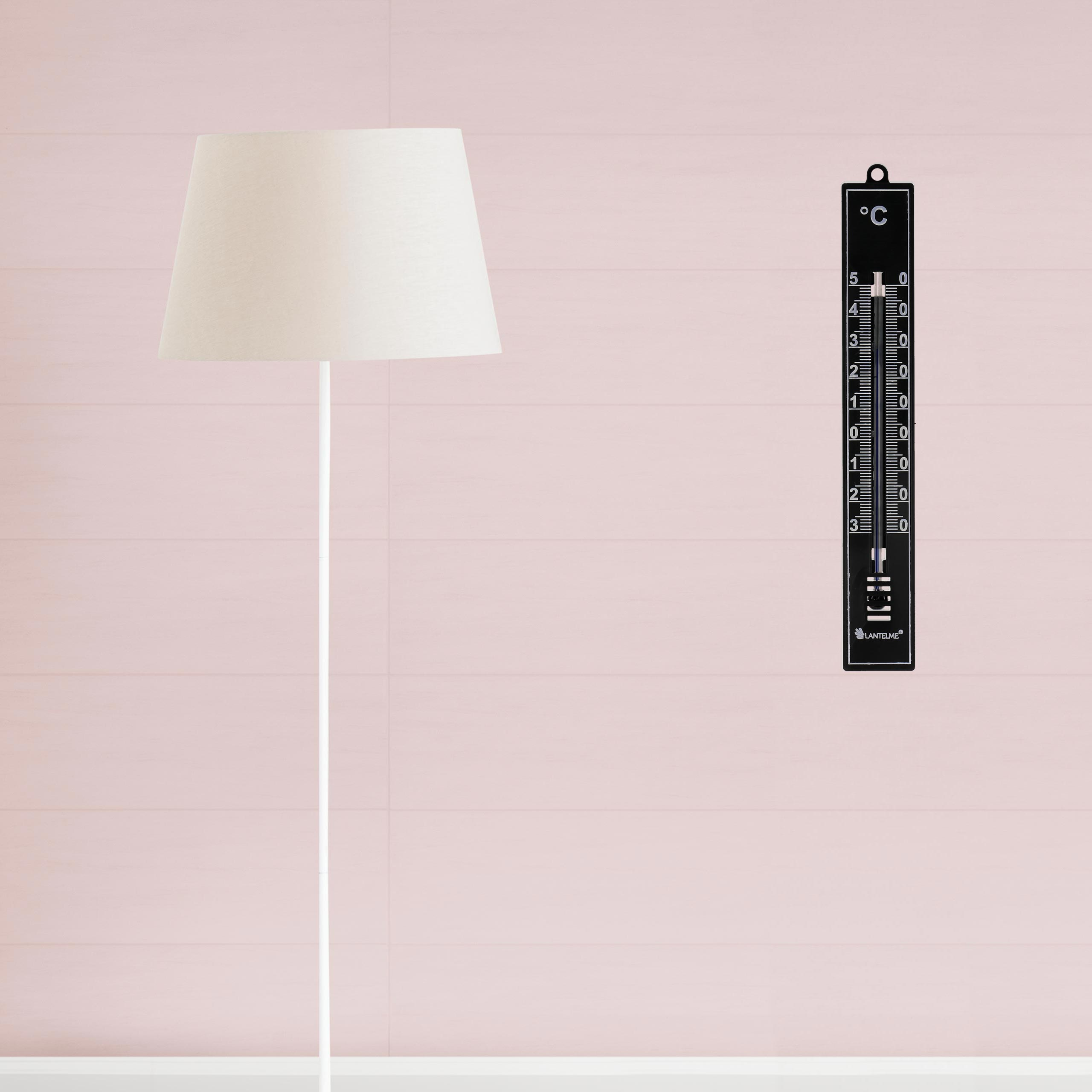 Ein schwarzes Thermometer an einer rosa Wand. Links ist eine weiße Lampe.