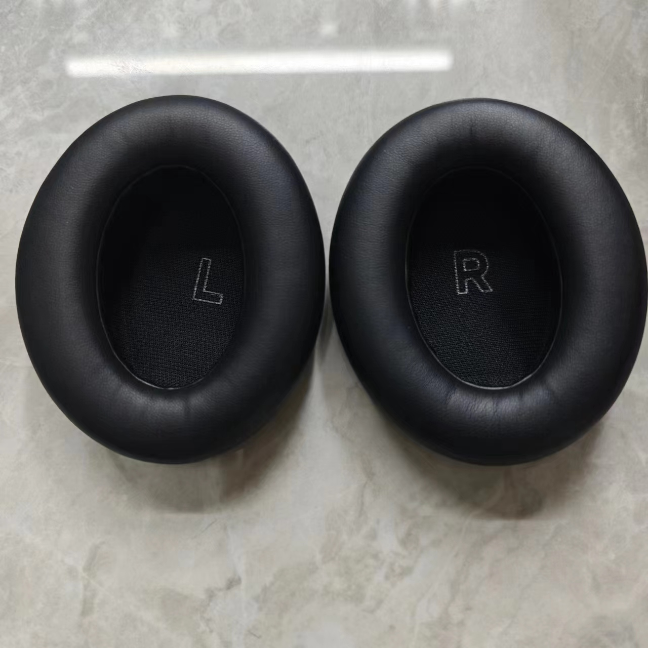 Twee zwarte oorkussens voor een koptelefoon, links met 'L', rechts met 'R', op een marmeren oppervlak.