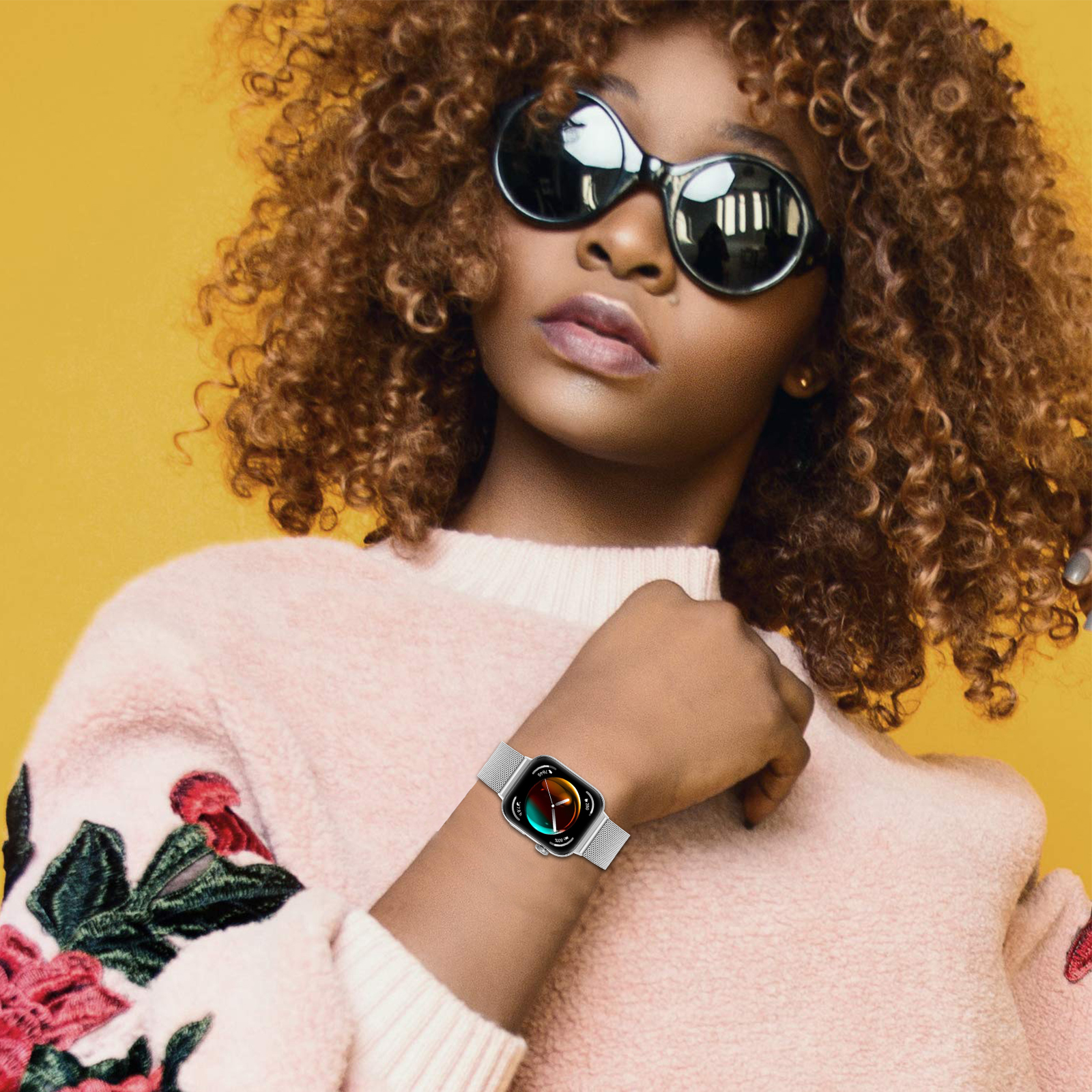 Kobieta w okularach przeciwsłonecznych i zegarku. Żółte tło. Ma na sobie różowy sweter.