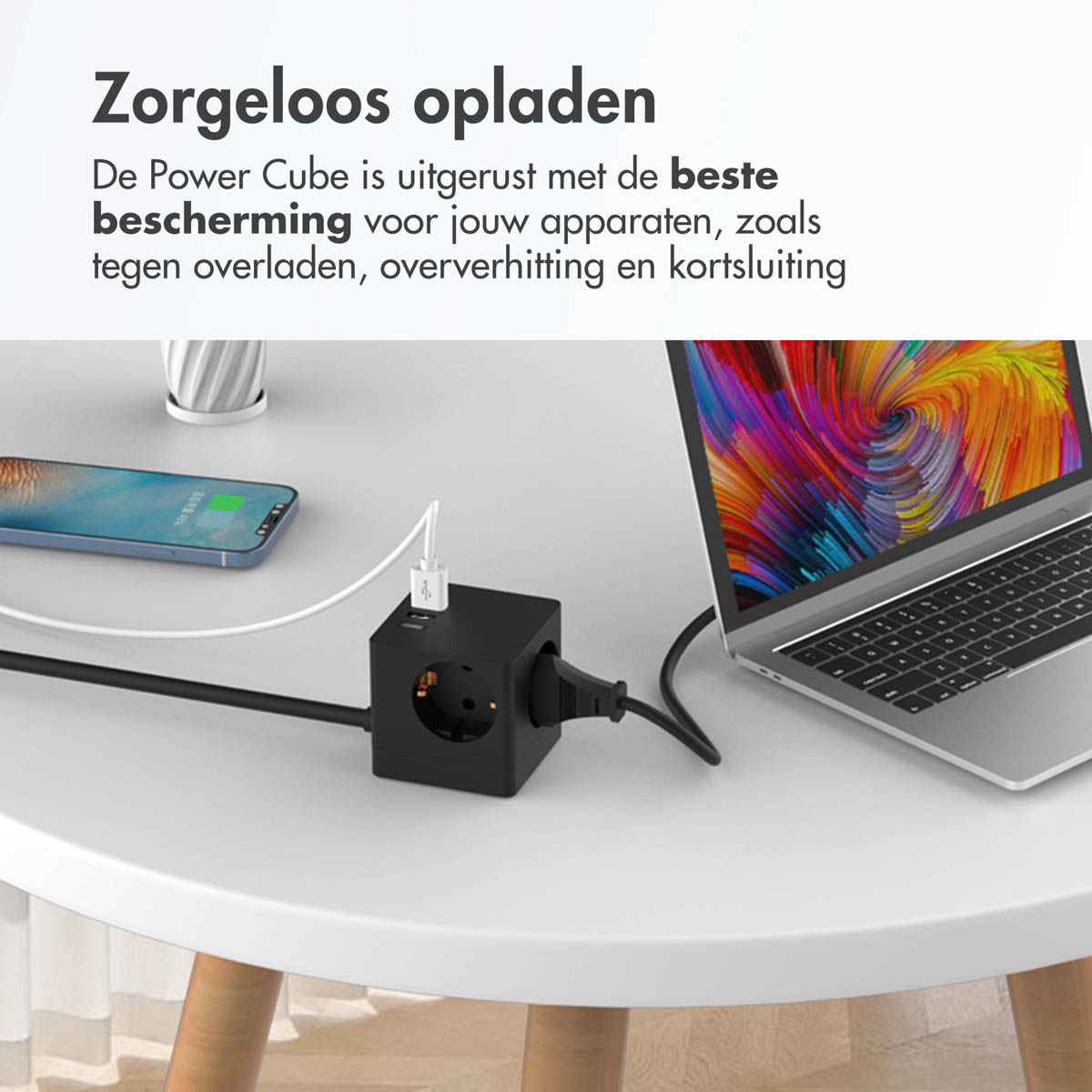 Een zwarte power cube met telefoon en laptop op een witte tafel. Zwart netsnoer zichtbaar.