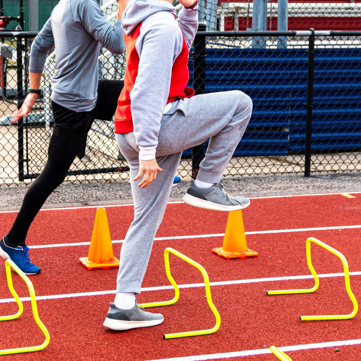 Two athletes run on a track. One steps on a cone, others jump over hurdles.