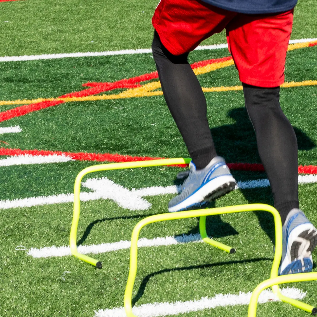 Athlete in red shorts running over yellow hurdles on a green track field.