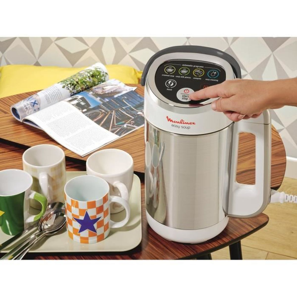 Robot da cucina, mano preme pulsante. Tazze e rivista vicine.