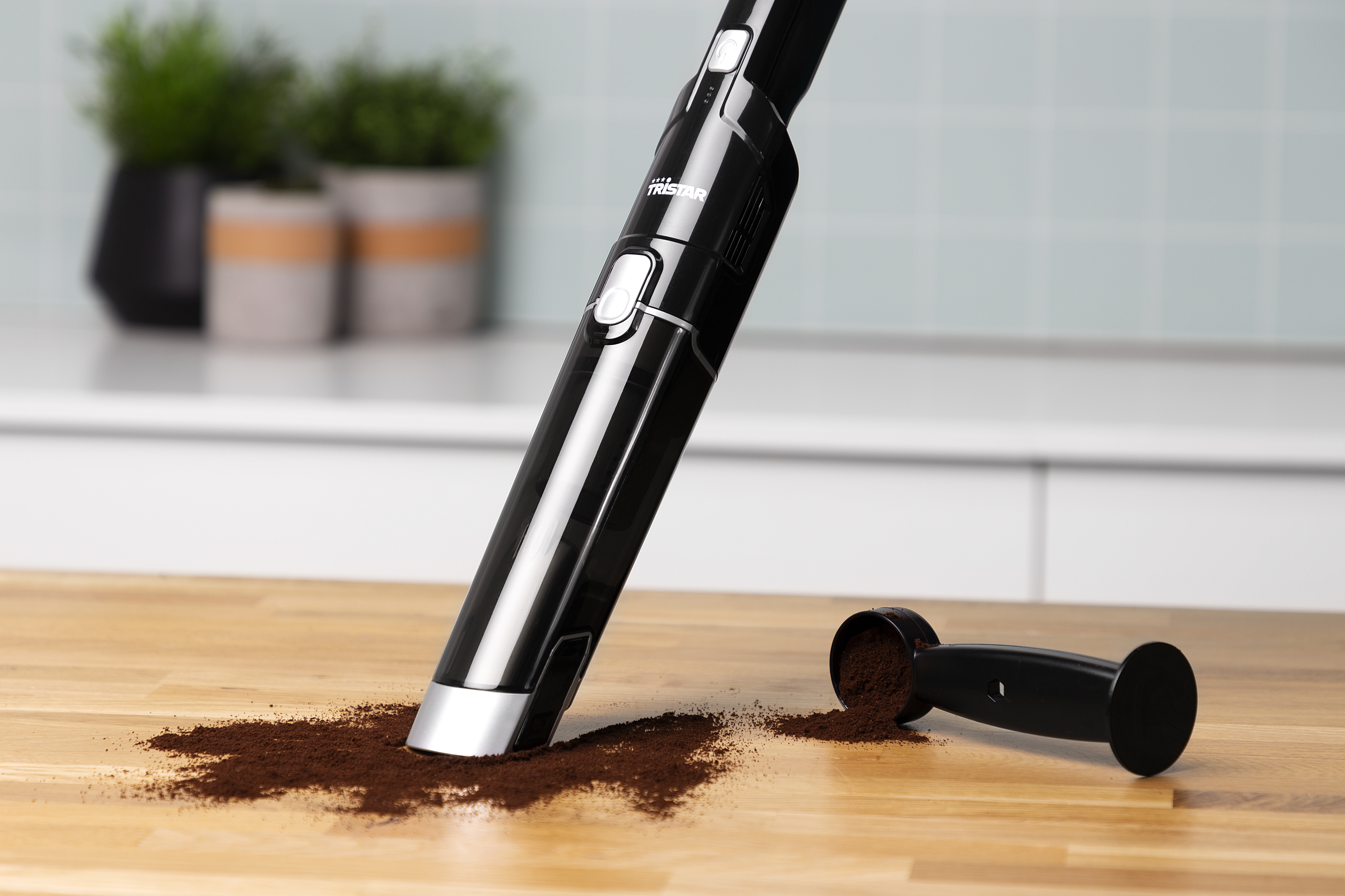 Un aspirapolvere portatile nero aspira fondi di caffè rovesciati su una superficie di legno.