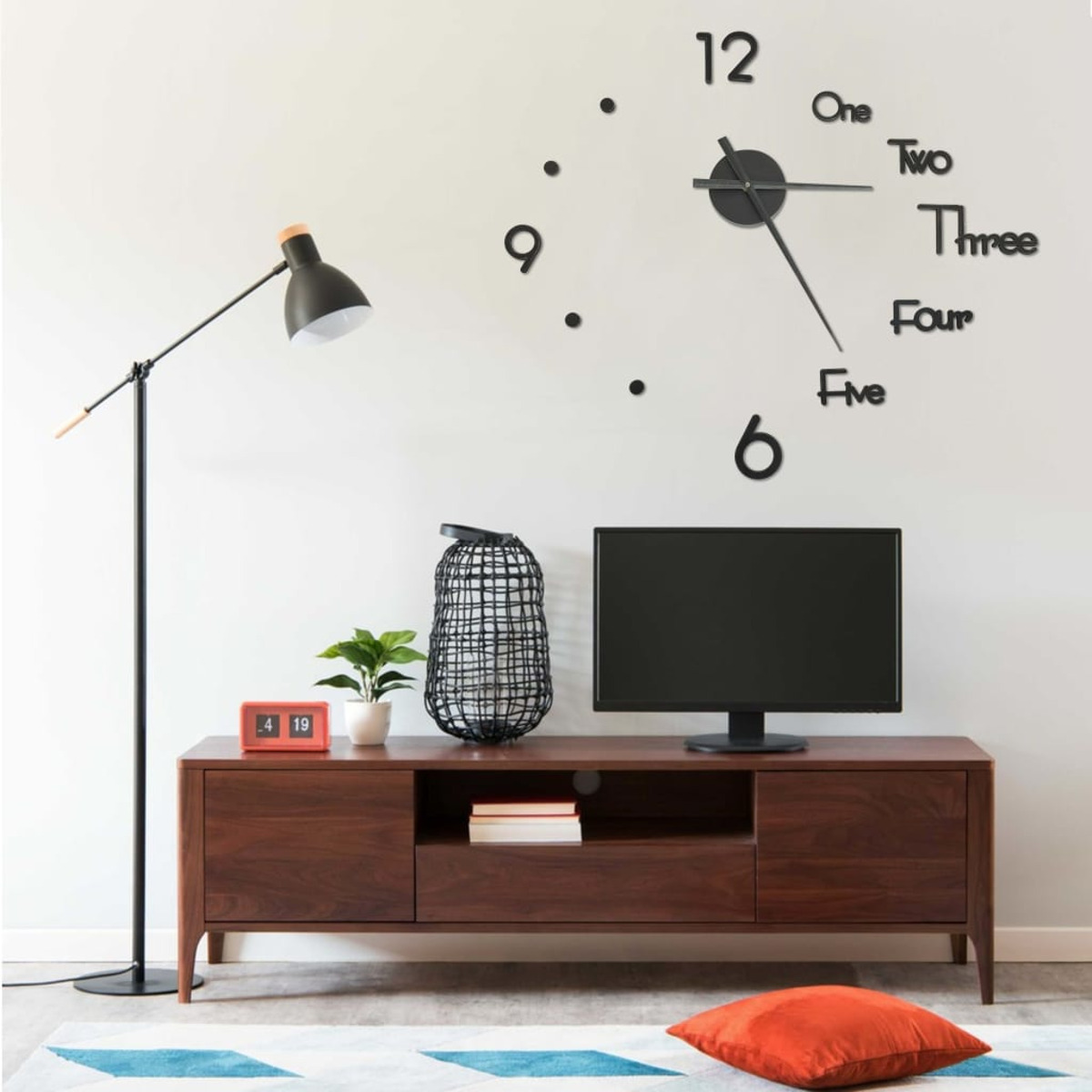 Wohnzimmer mit Uhr, Fernseher und Lampe. Ein Schrank unter dem Fernseher, rotes Kissen.