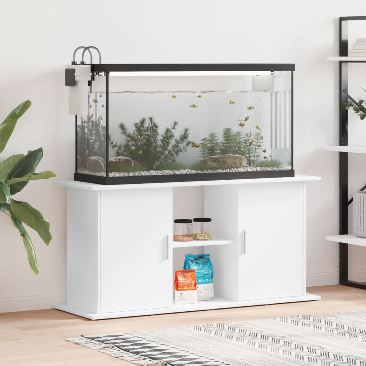 Ein weißes Aquarium auf einem weißen Schrank mit Futtersäcken und einer Pflanze im Hintergrund. Dekorationen sind vorhanden.