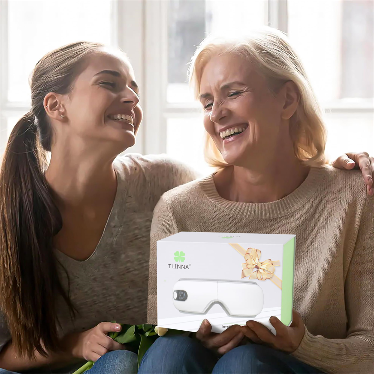 Zwei lächelnde Frauen halten eine Geschenkbox für ein Augenmassagegerät. Die Box hat ein Kleeblatt-Logo und eine Schleife.