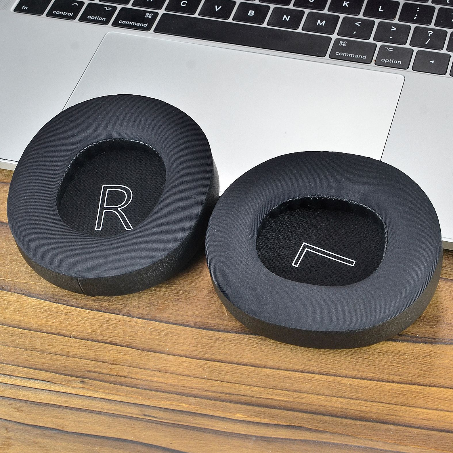 Dwie czarne poduszki nauszne słuchawek z literami R i L. Leżą na drewnianej powierzchni.