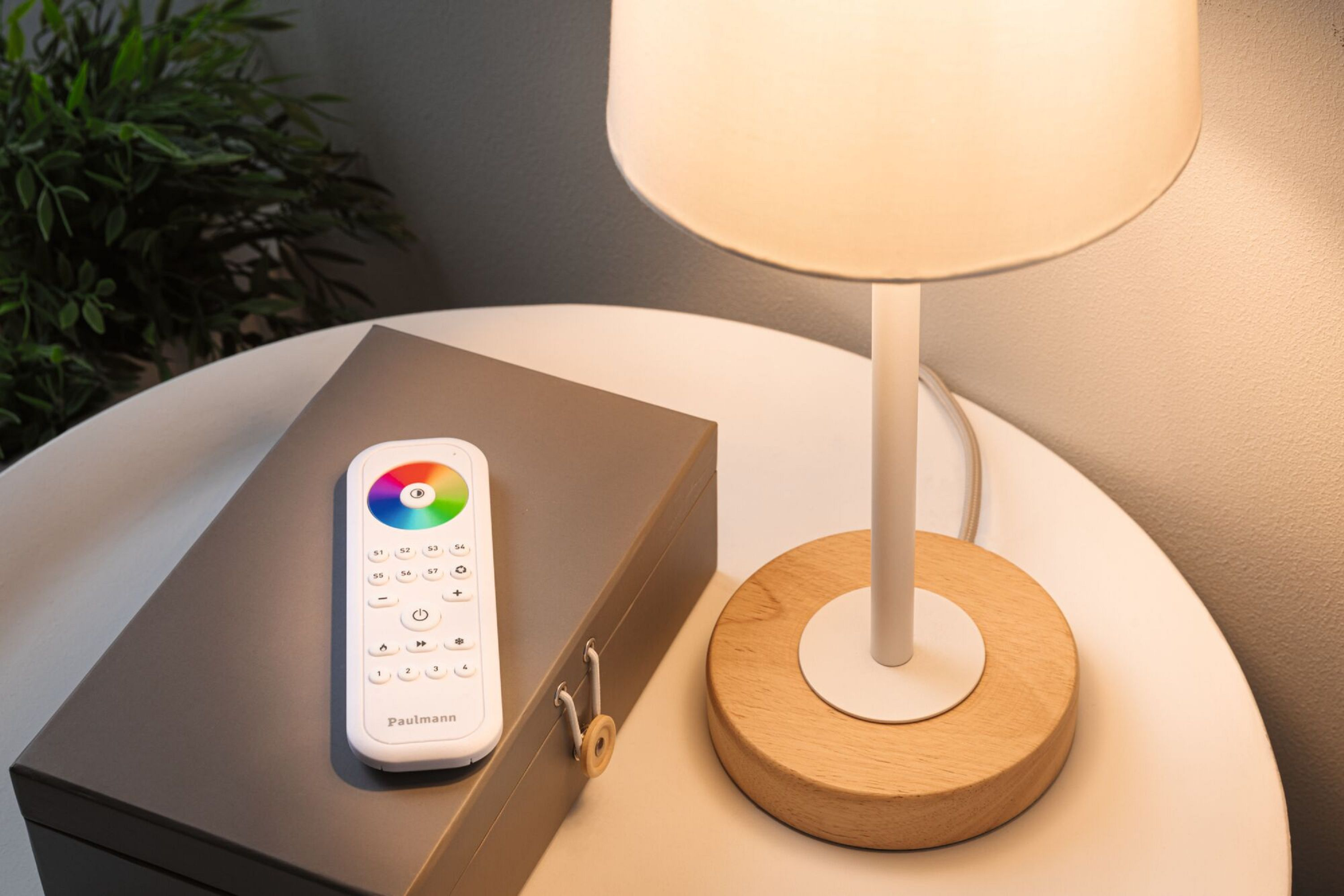 Tischlampe mit weißem Lampenschirm und Holzsockel. Fernbedienung und graue Box auf dem Tisch.