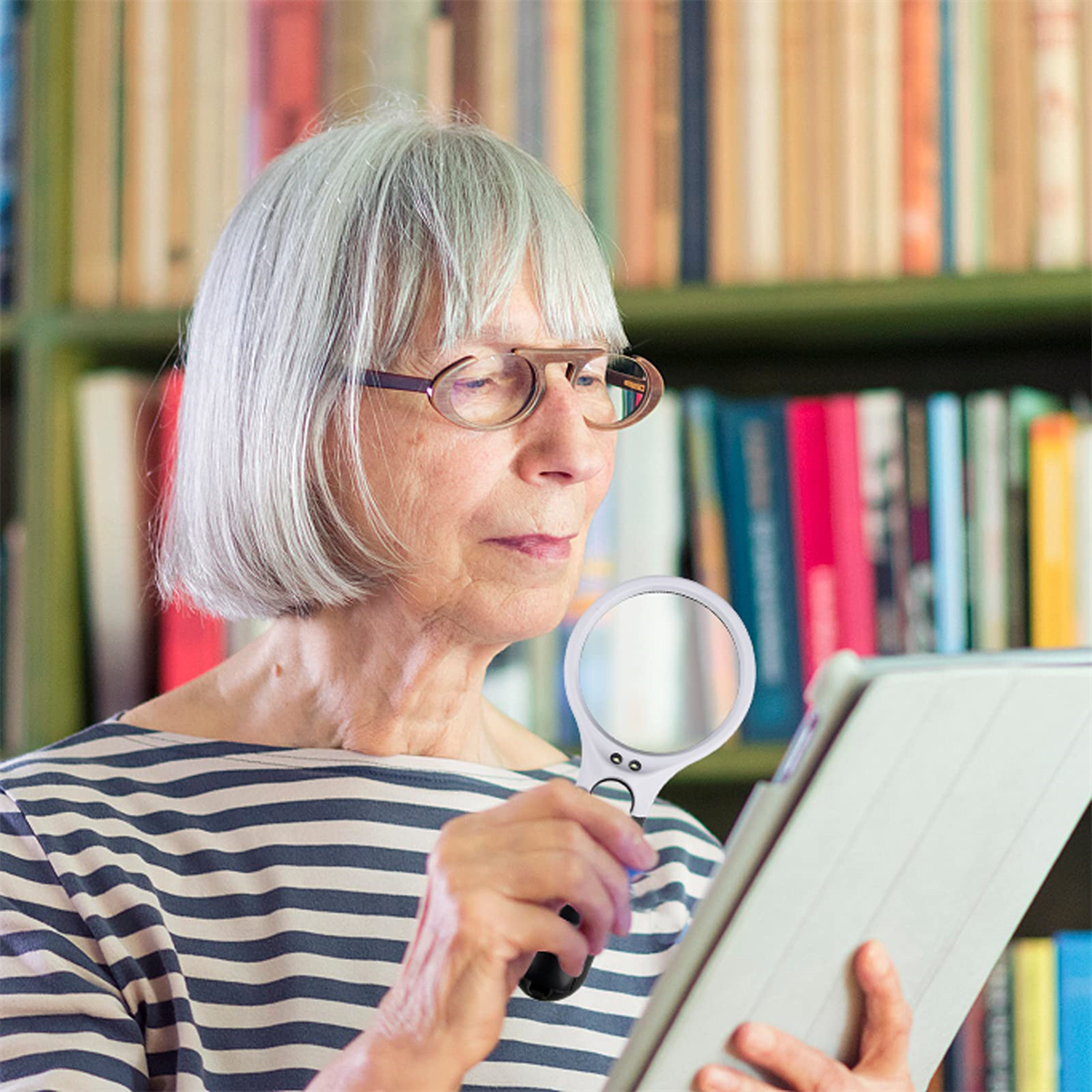 Eine ältere Frau mit Brille liest mit einer Lupe ein Tablet vor Bücherregalen.