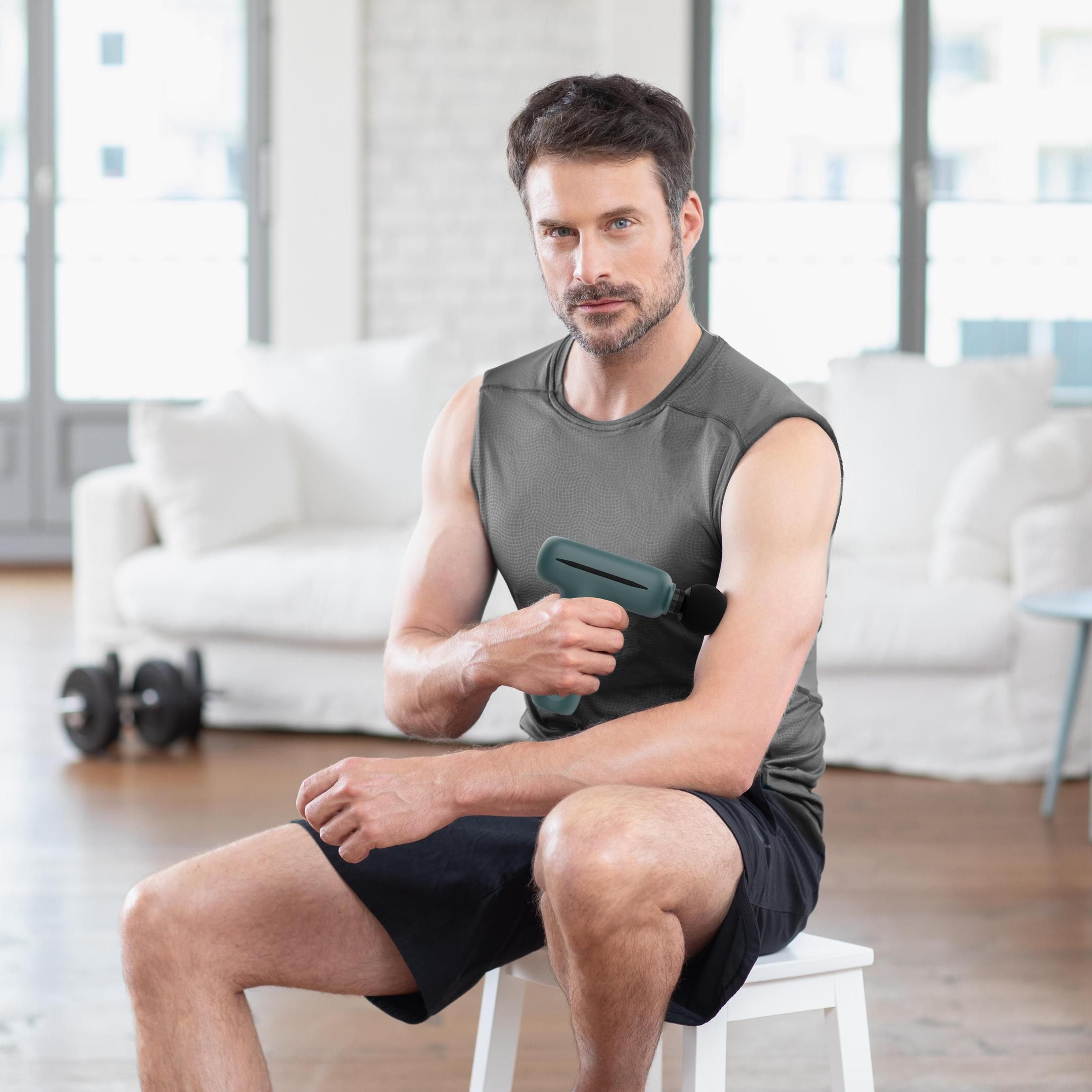 Mężczyzna siedzi na stołku używając pistoletu do masażu na ramieniu. Tło: kanapa i okno.