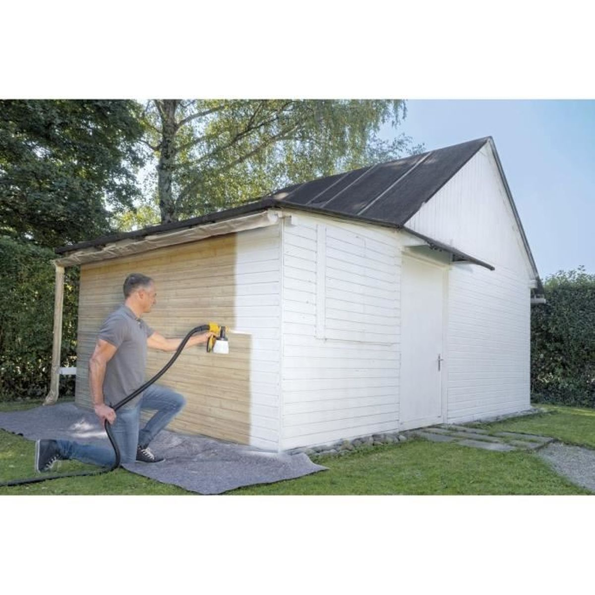 Ein Mann in Jeans streicht einen Holzschuppen mit einem Farbsprühgerät weiß an. Der Boden ist bedeckt.