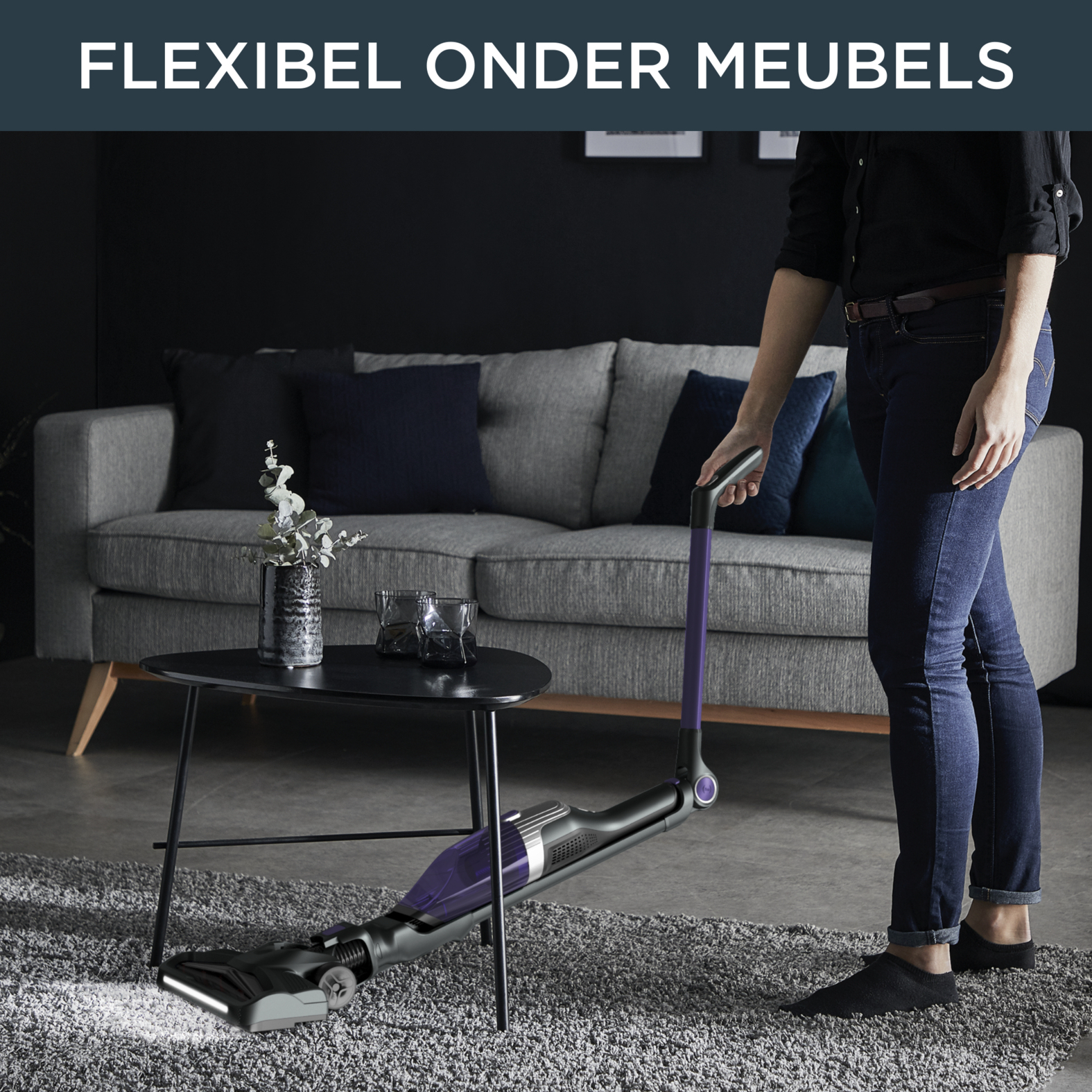 Vrouw die onder een tafel stofzuigt. Donkergrijs tapijt en een bank. Tekst: Flexibel onder meubels.