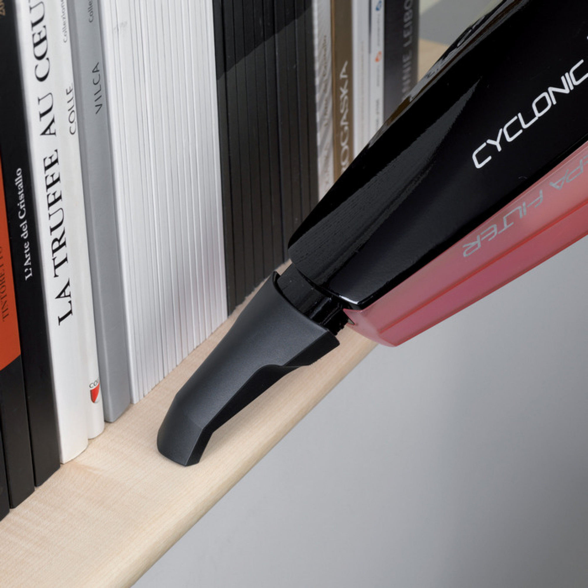Ein schwarz-roter Handstaubsauger reinigt ein Regal mit Büchern. Der Staubsauger berührt das Regal.