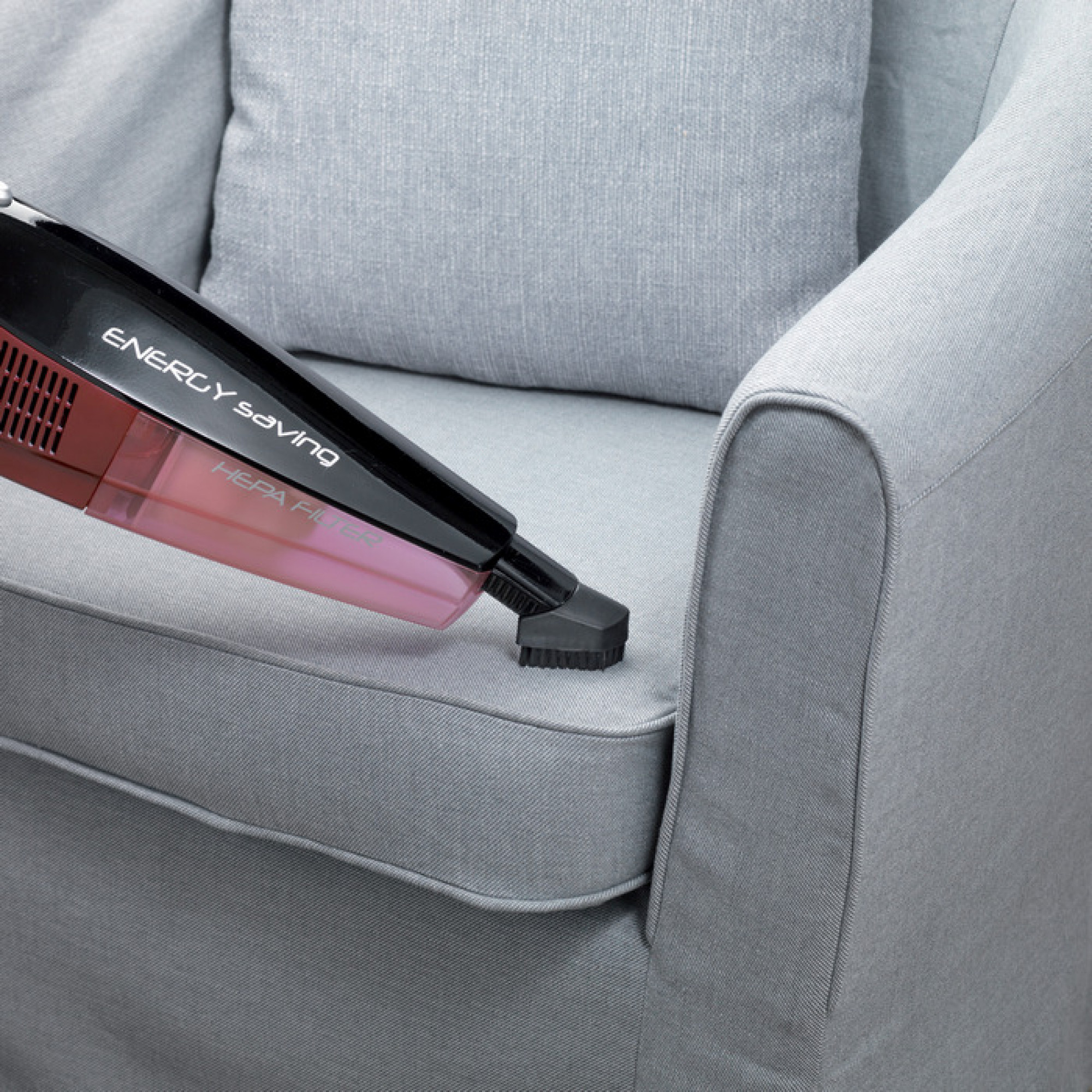 Ein Handstaubsauger reinigt ein graues Sofa. Der Staubsauger ist schwarz-rot und hat einen HEPA-Filter.