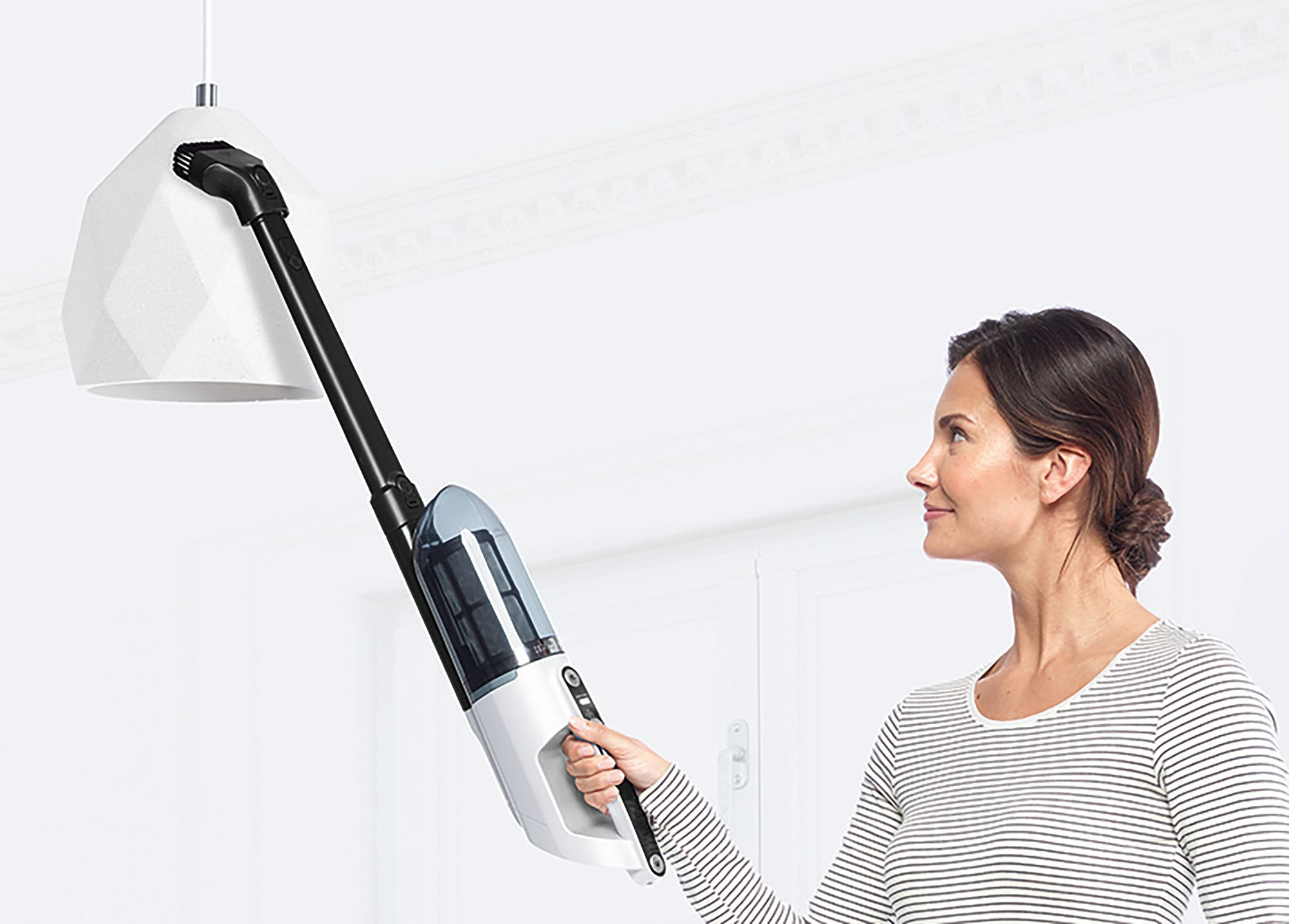 Frau reinigt mit einem kabellosen Staubsauger eine weiße Lampe und blickt zum Licht.