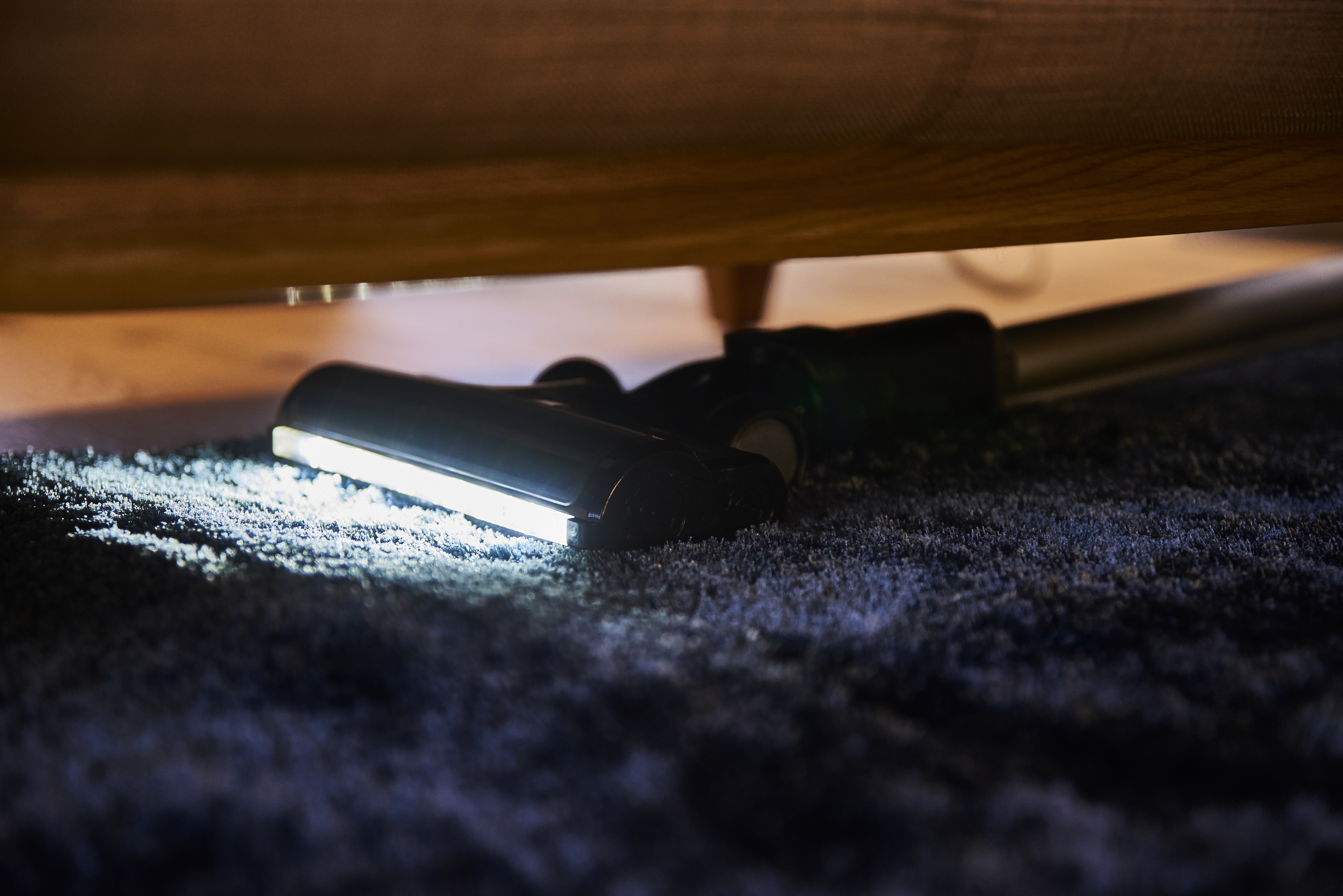 Testa dell'aspirapolvere sotto i mobili, illuminata. Tappeto grigio scuro. Gambe in legno visibili.