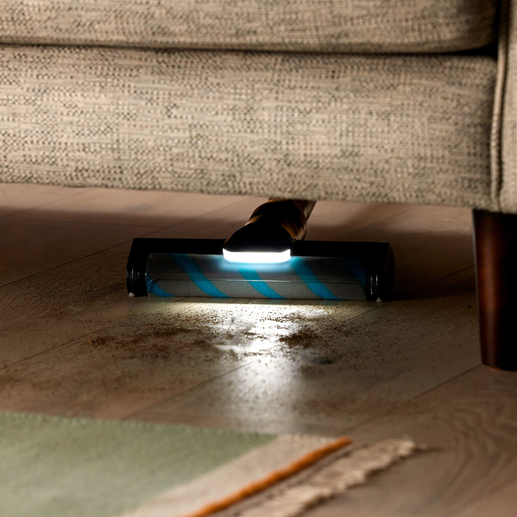 Staubsaugerkopf reinigt den Boden unter einem Sofa, beleuchtet durch blaues Licht.