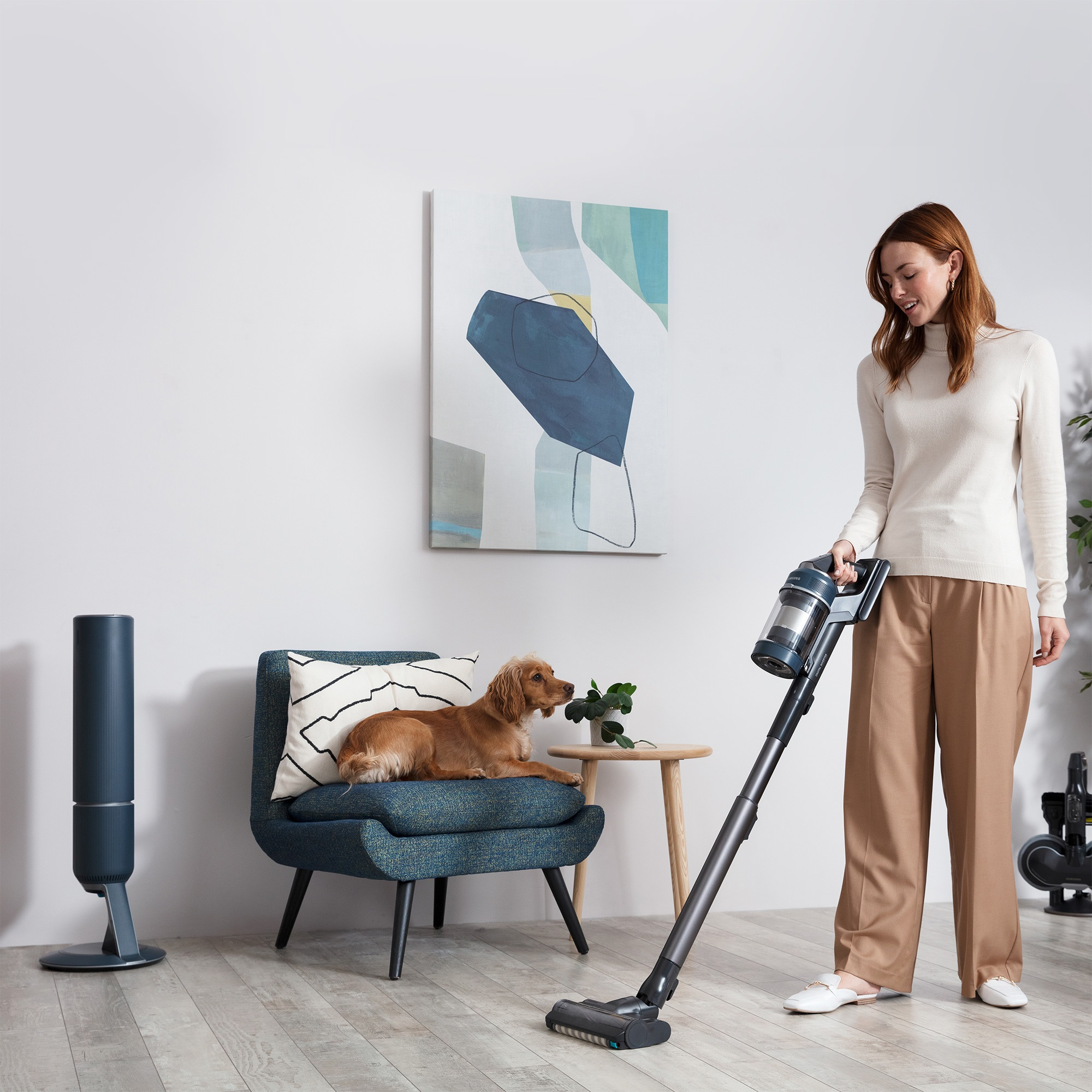 Eine Frau saugt mit einem kabellosen Stielsauger, Hund auf Stuhl, Kunstwerk an der Wand.