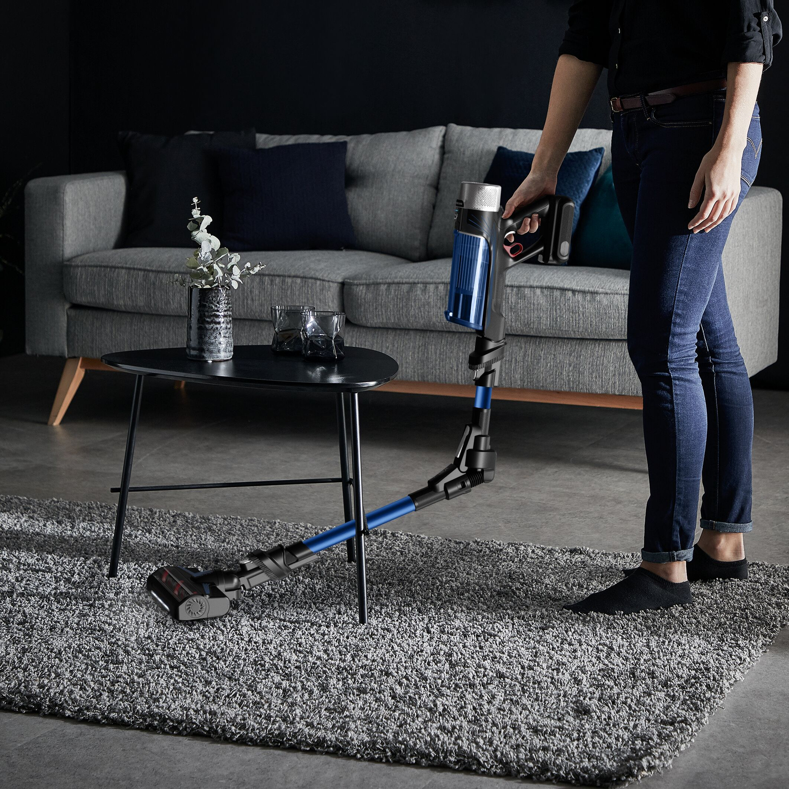 Una persona aspira una alfombra en una sala de estar. Se ven un sofá gris y una mesa auxiliar negra.