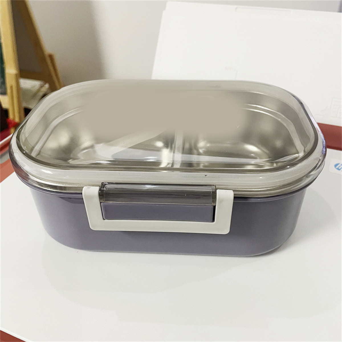 Eine grau-violette Lunchbox mit durchsichtigem Deckel liegt auf einer weißen Oberfläche.