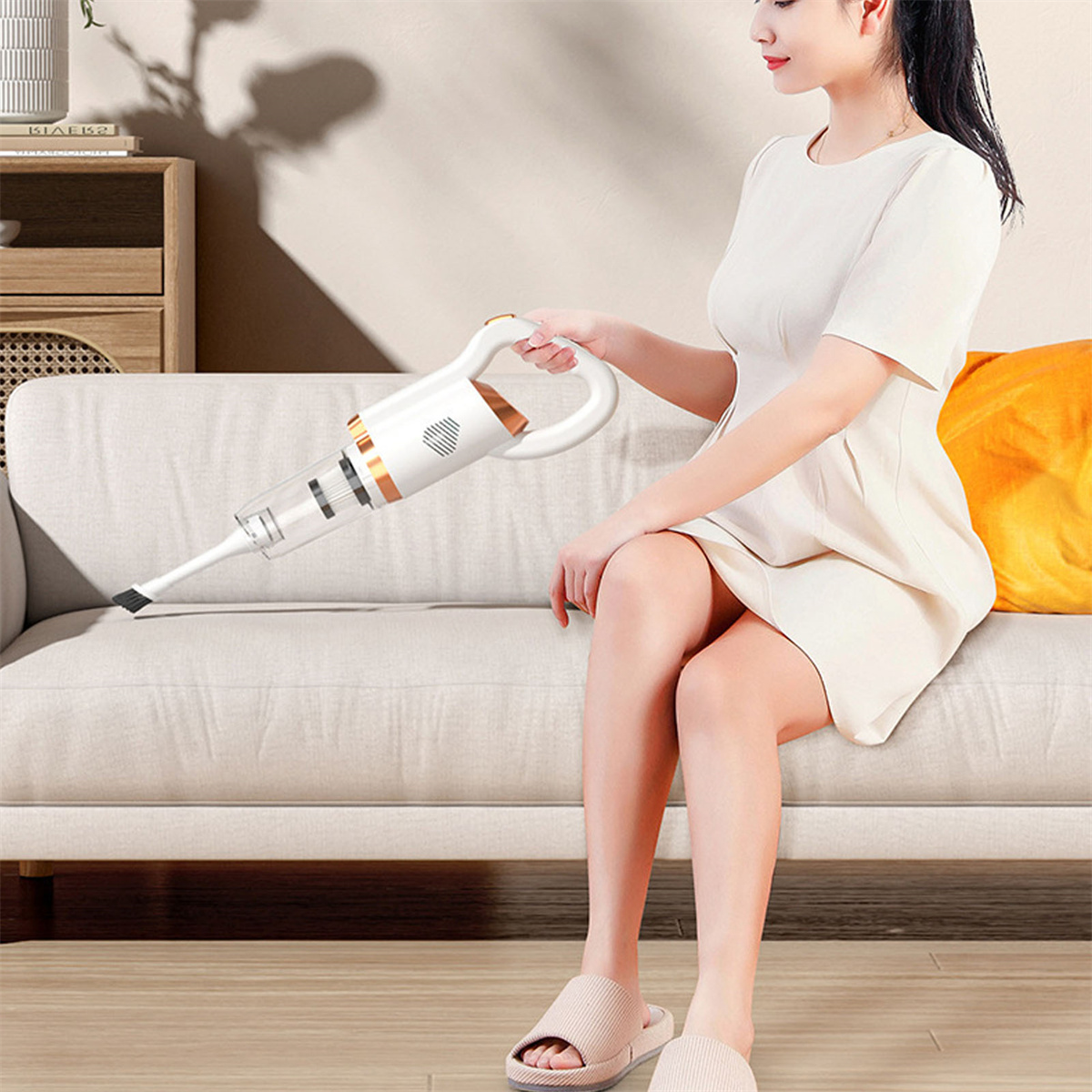 Eine Frau benutzt einen Handstaubsauger auf einem weißen Sofa. Beige Hausschuhe und orangefarbenes Kissen.