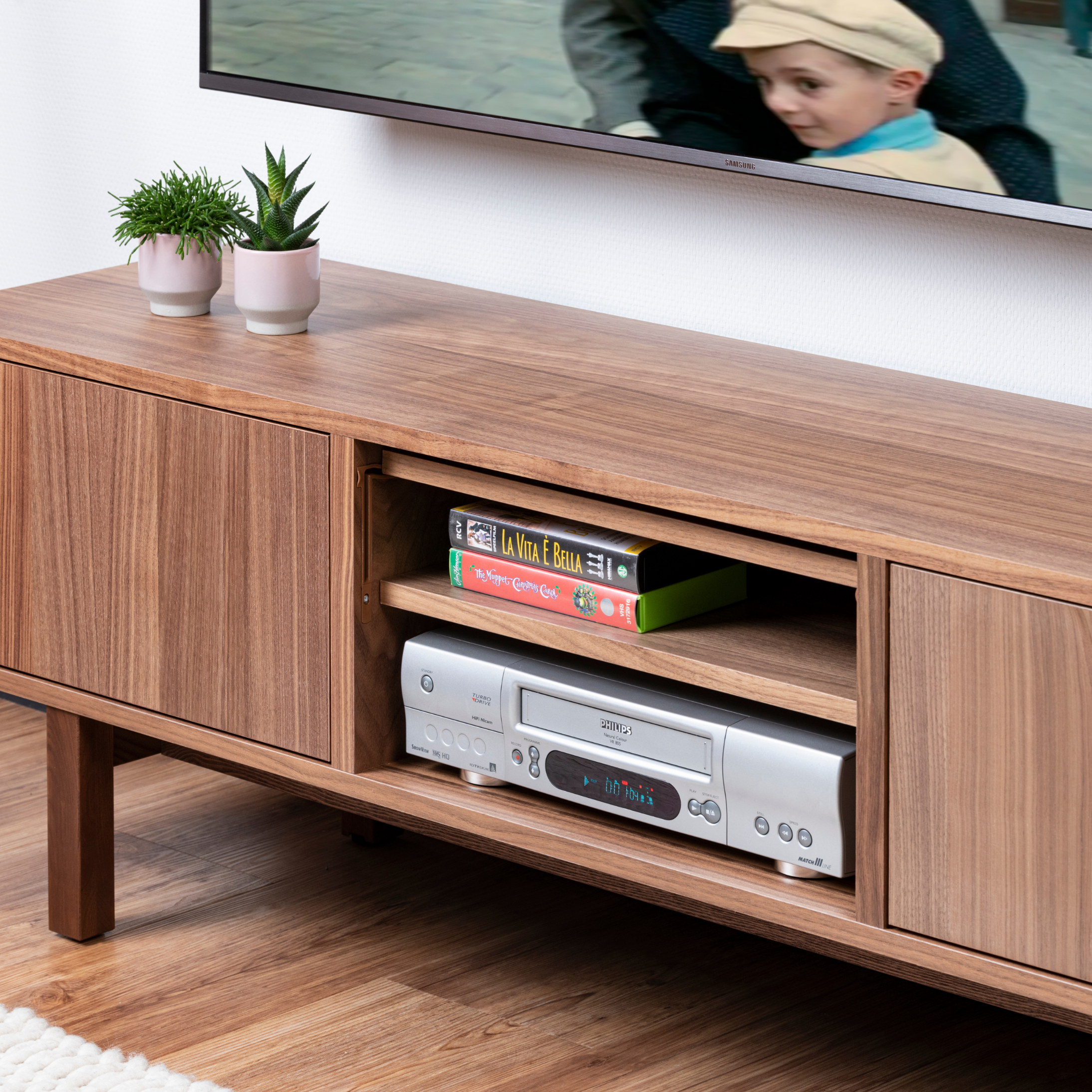 TV-Ständer aus Holz mit einem VCR und einem Film. Ein Fernseher befindet sich über dem Ständer. Pflanzen.