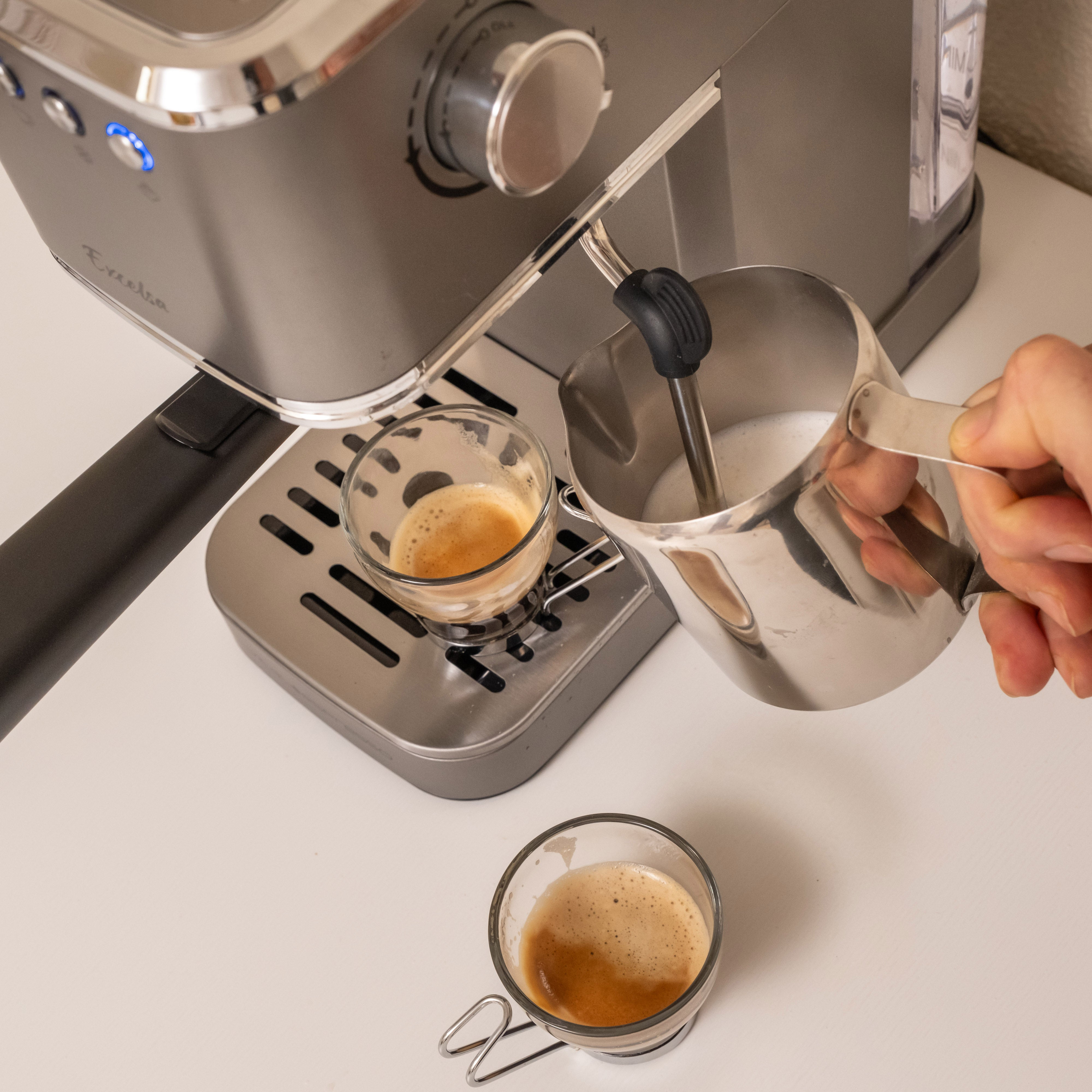 Una persona vierte leche en dos tazas de espresso. La máquina de espresso es gris.