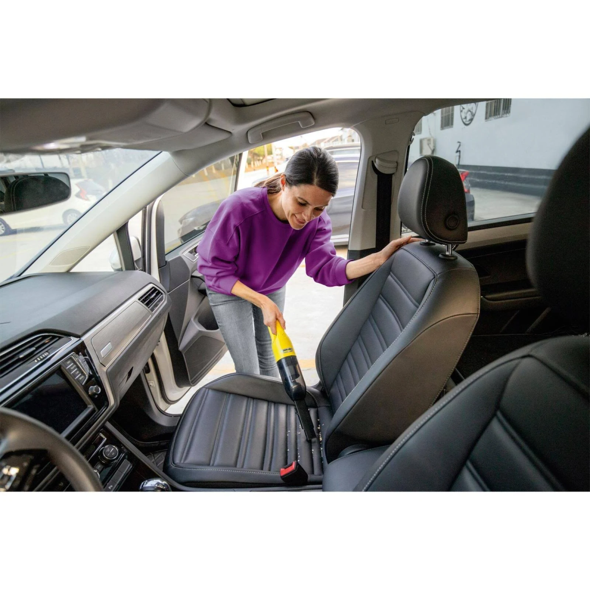 Vrouw stofzuigt een autostoel met een gele en zwarte handstofzuiger. Achtergrond toont het interieur van de auto.