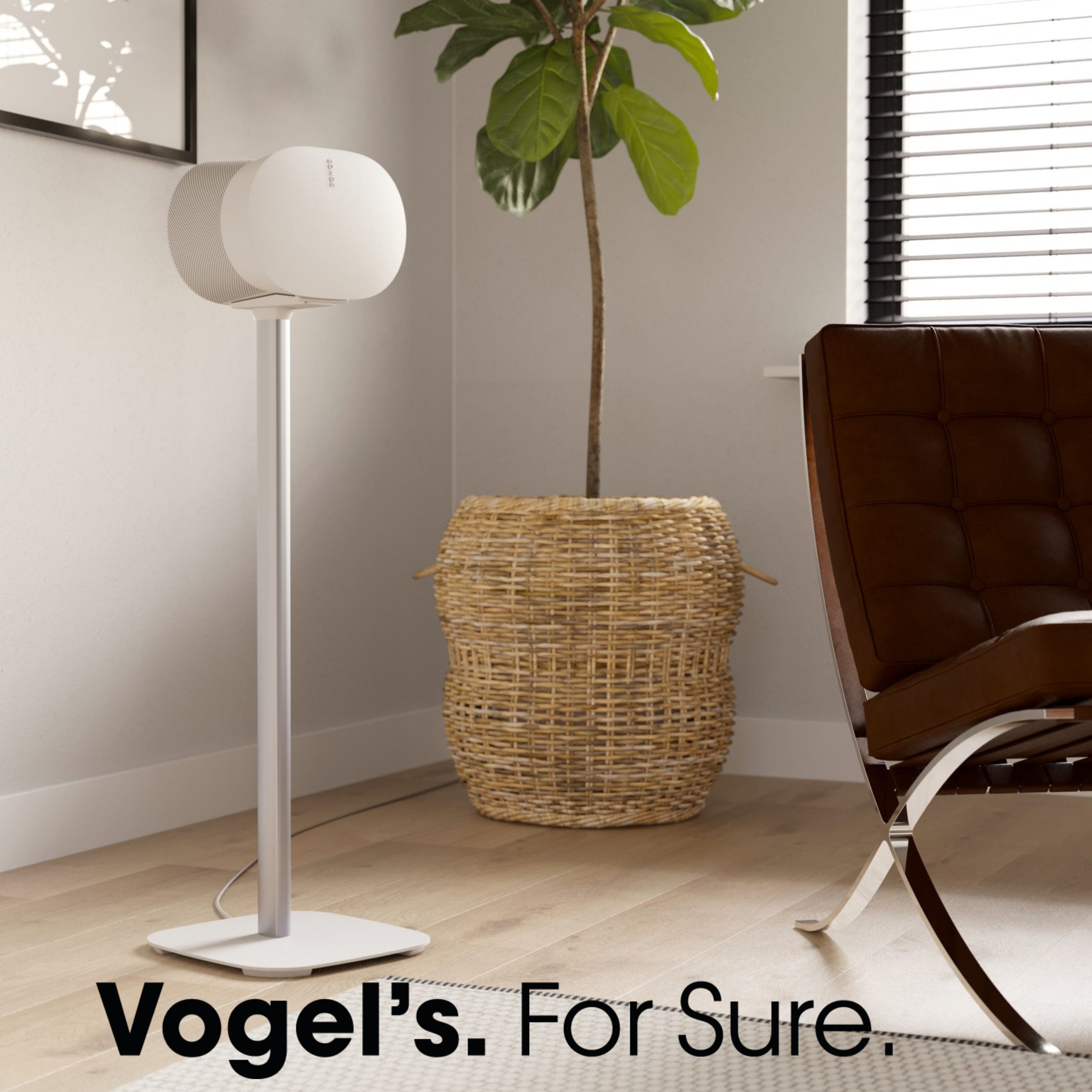 Biały głośnik na stojaku obok rośliny doniczkowej i brązowego krzesła. Tekst: 'Vogel's. For Sure'.