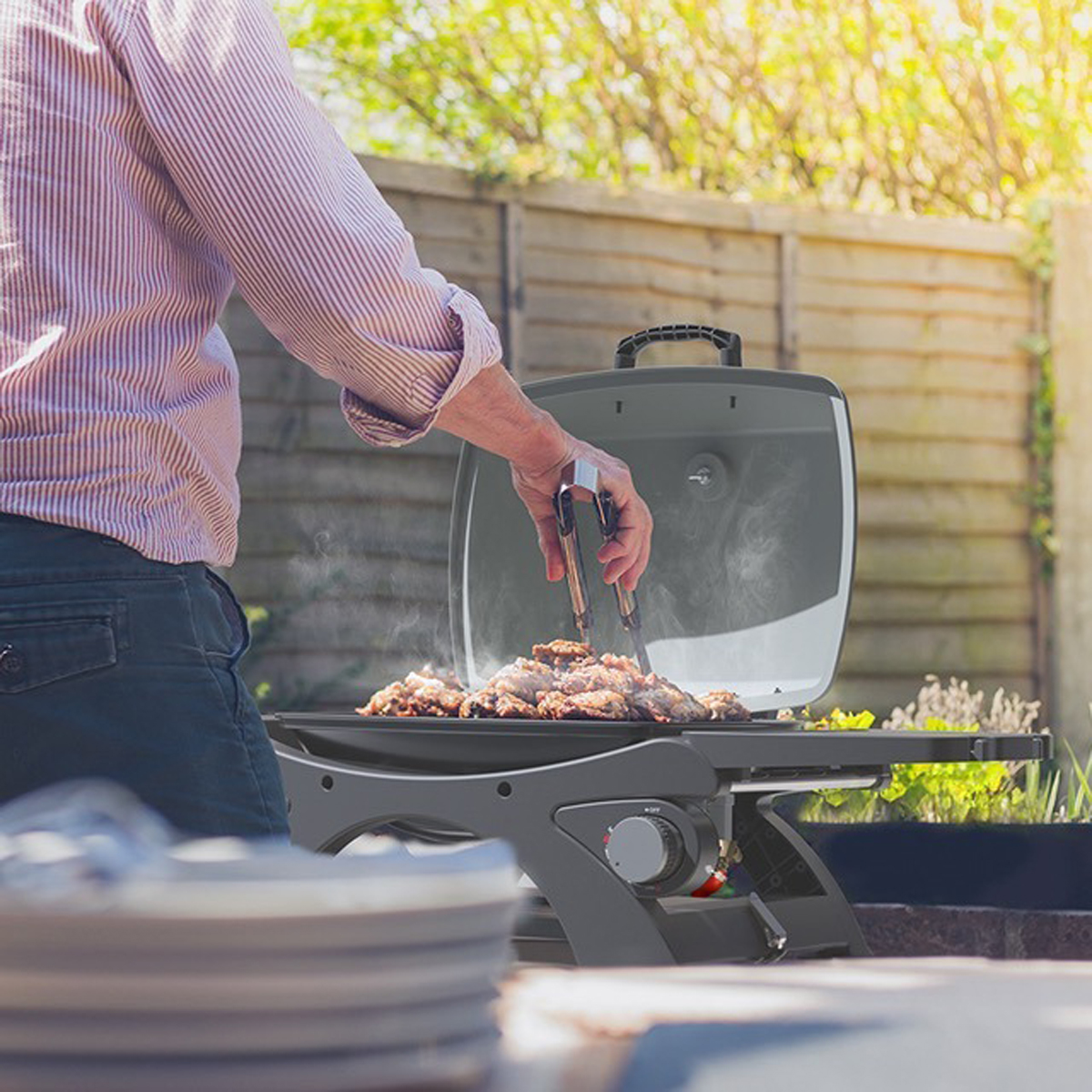 Personne grillant des aliments sur un barbecue gris Kitchen Brothers. La scène est en extérieur, avec une clôture en bois.