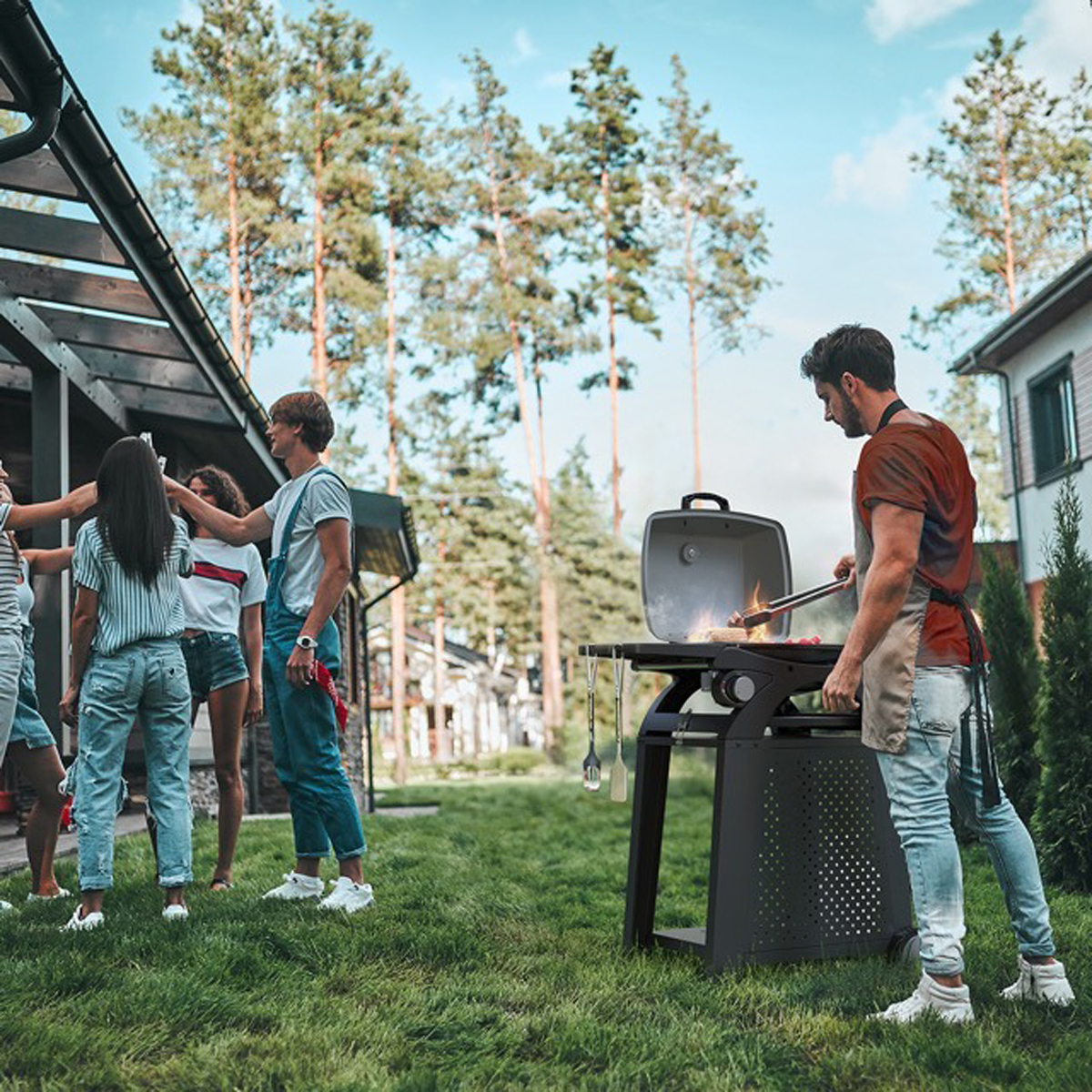 Des gens font des grillades sur un gril noir devant une maison. Un homme cuisine et d'autres personnes socialisent.