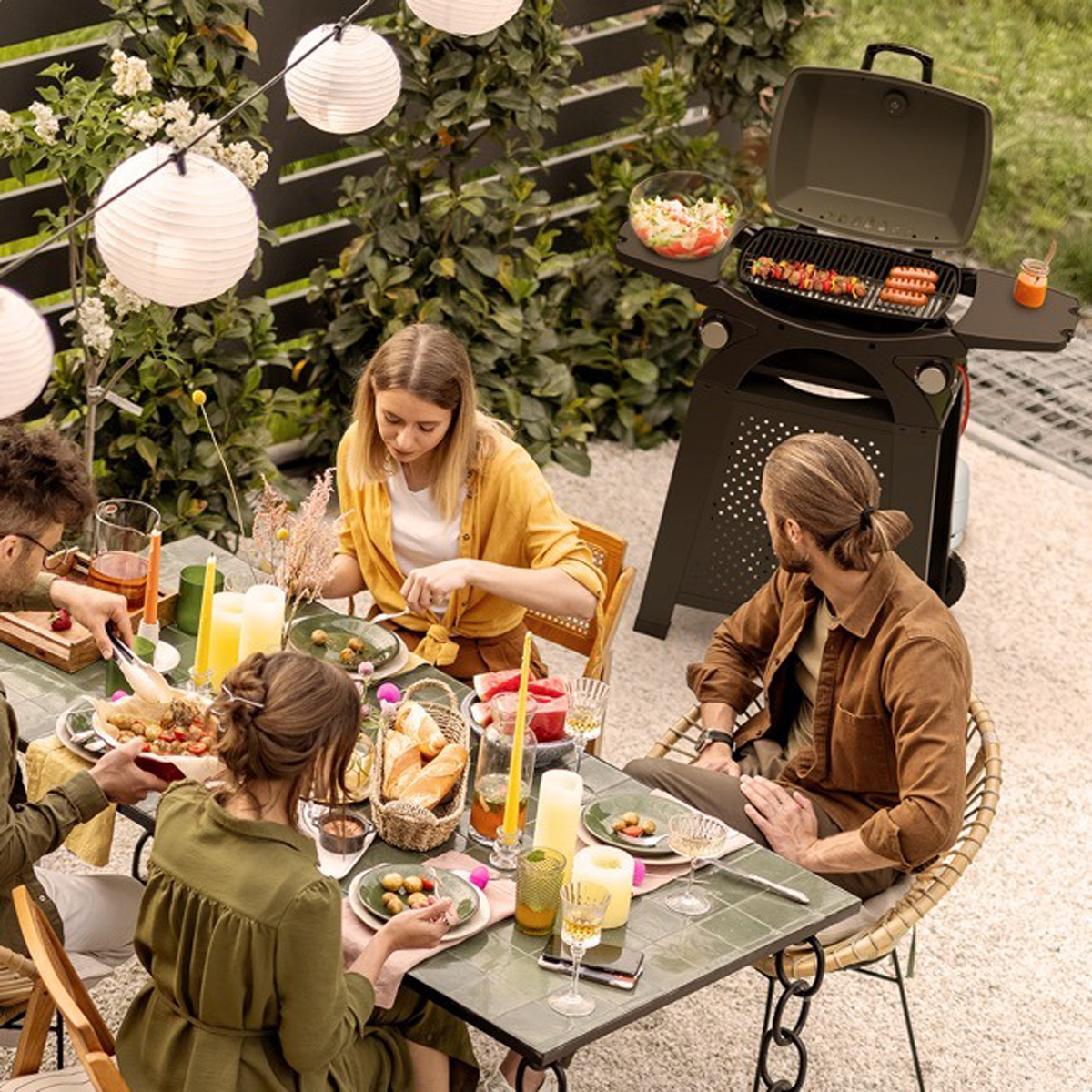 Un groupe de personnes à une table profitant d'un barbecue. Un grill est à l'arrière-plan. La scène est en plein air.