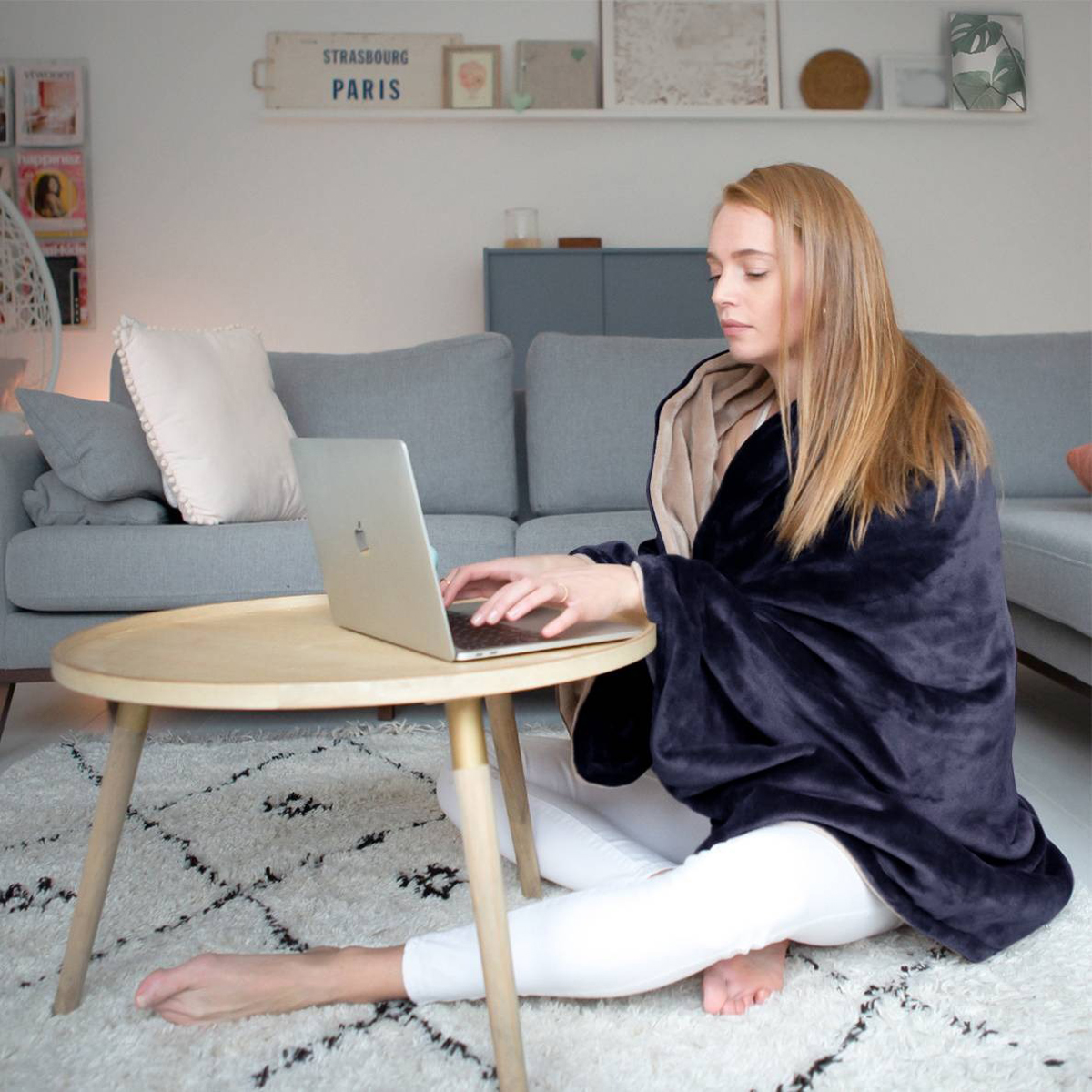 Woman using a laptop, wrapped in a dark blue blanket, sitting on a rug by a coffee table.