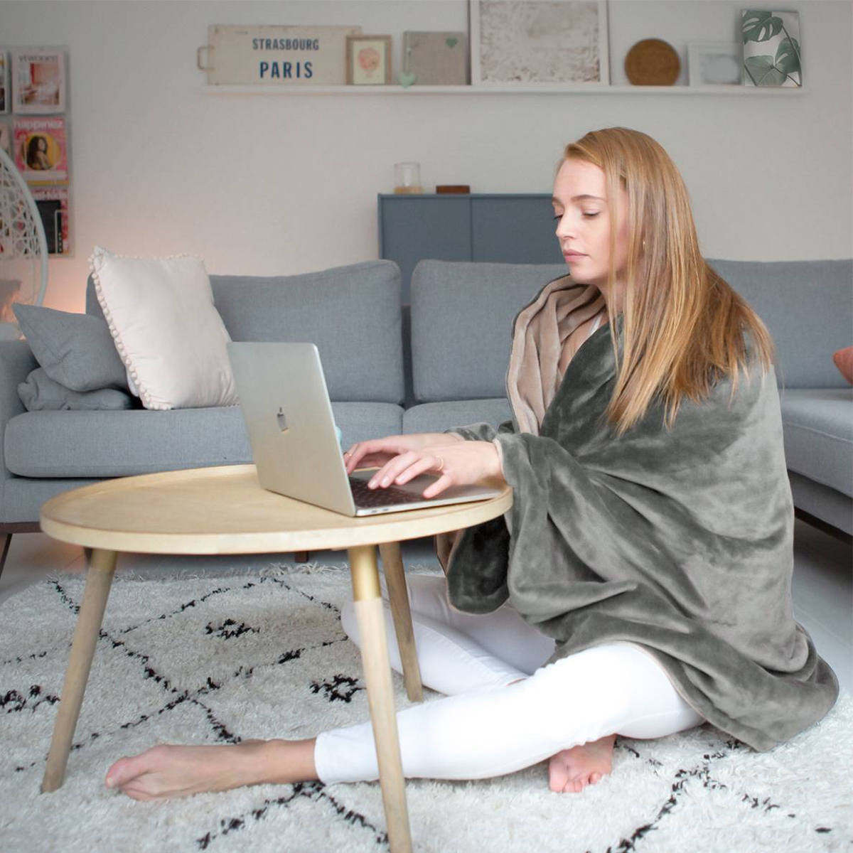 Woman in a living room using a laptop, wrapped in a blanket, sitting near a wooden table.