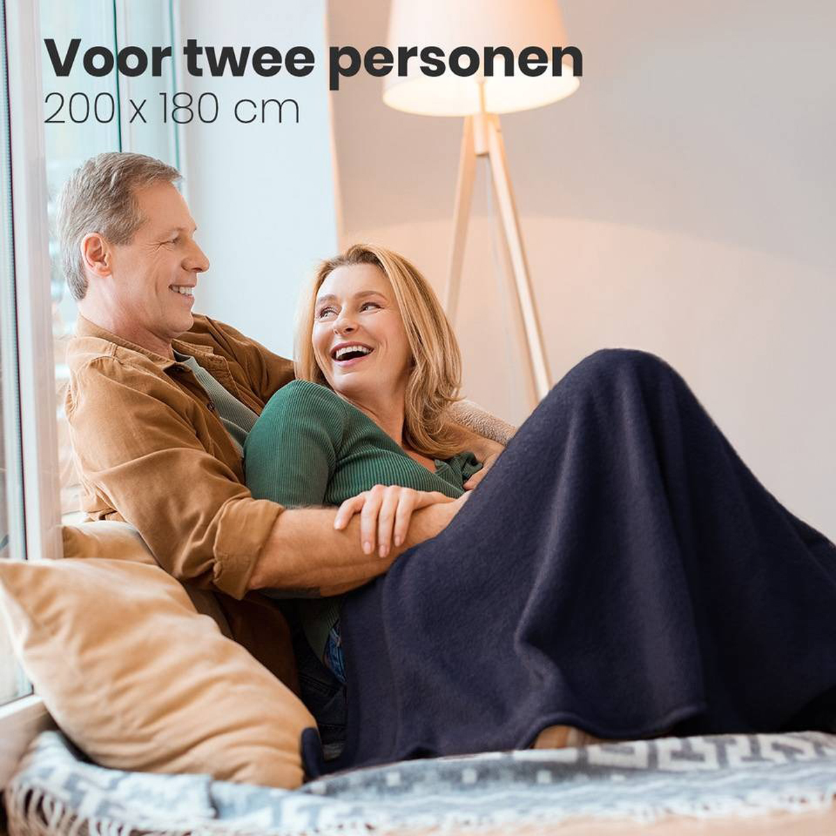 Couple sitting, wrapped in a blanket. Smiling in a room with a lamp and a window.