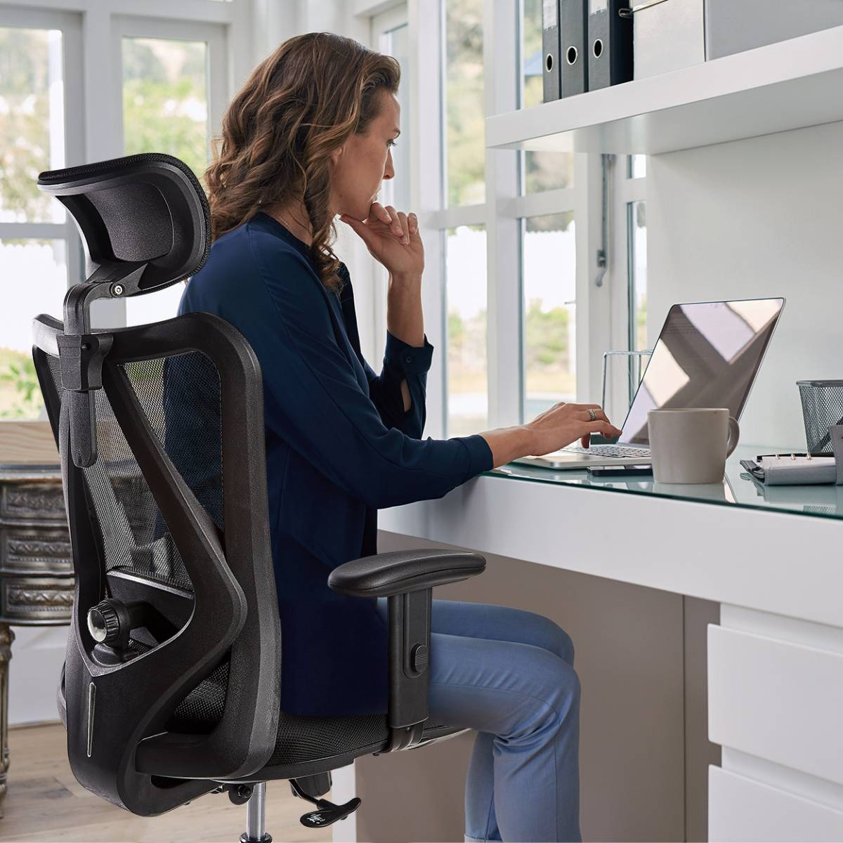 A woman is seated at a desk using a laptop, with a black office chair behind her.